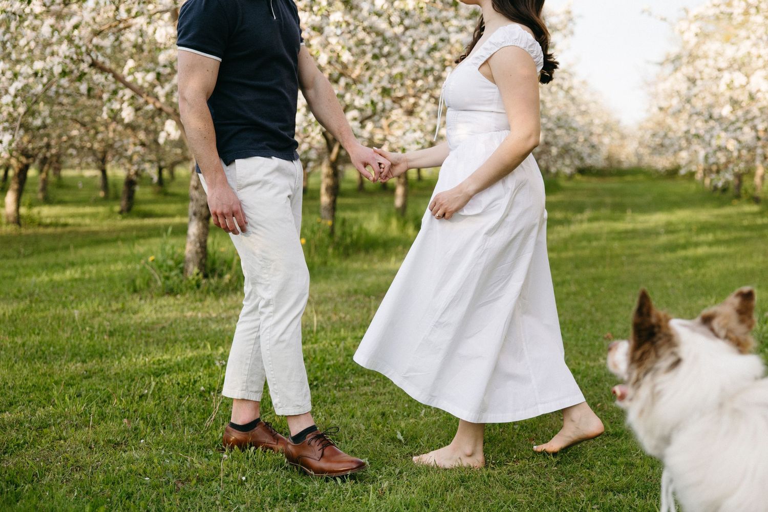 Pregnant woman in white dress holds hands with man in black shirt among flowering trees barefoot.