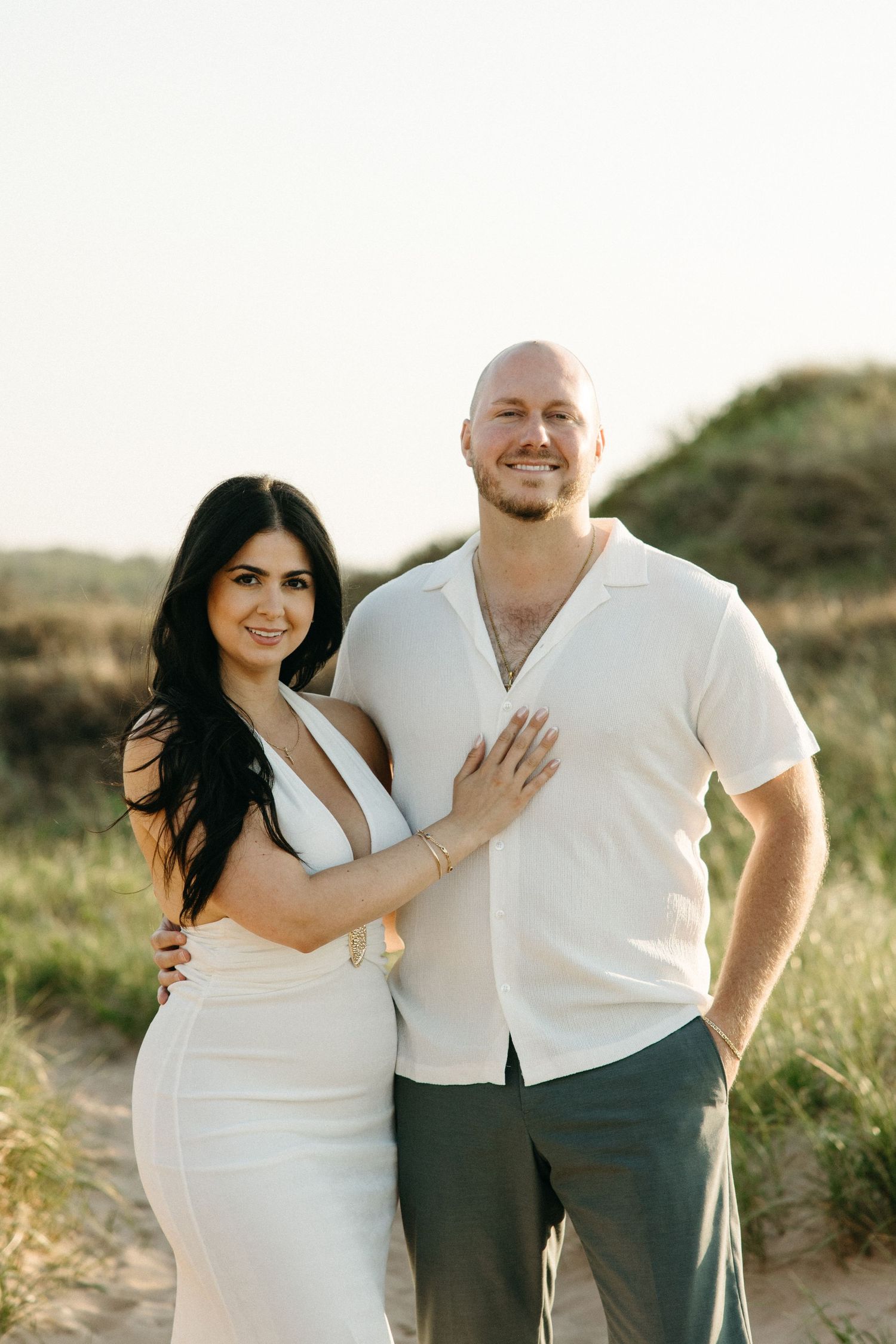 A smiling couple poses on sandy beach dunes, woman in white dress, man in white shirt and gray pants.