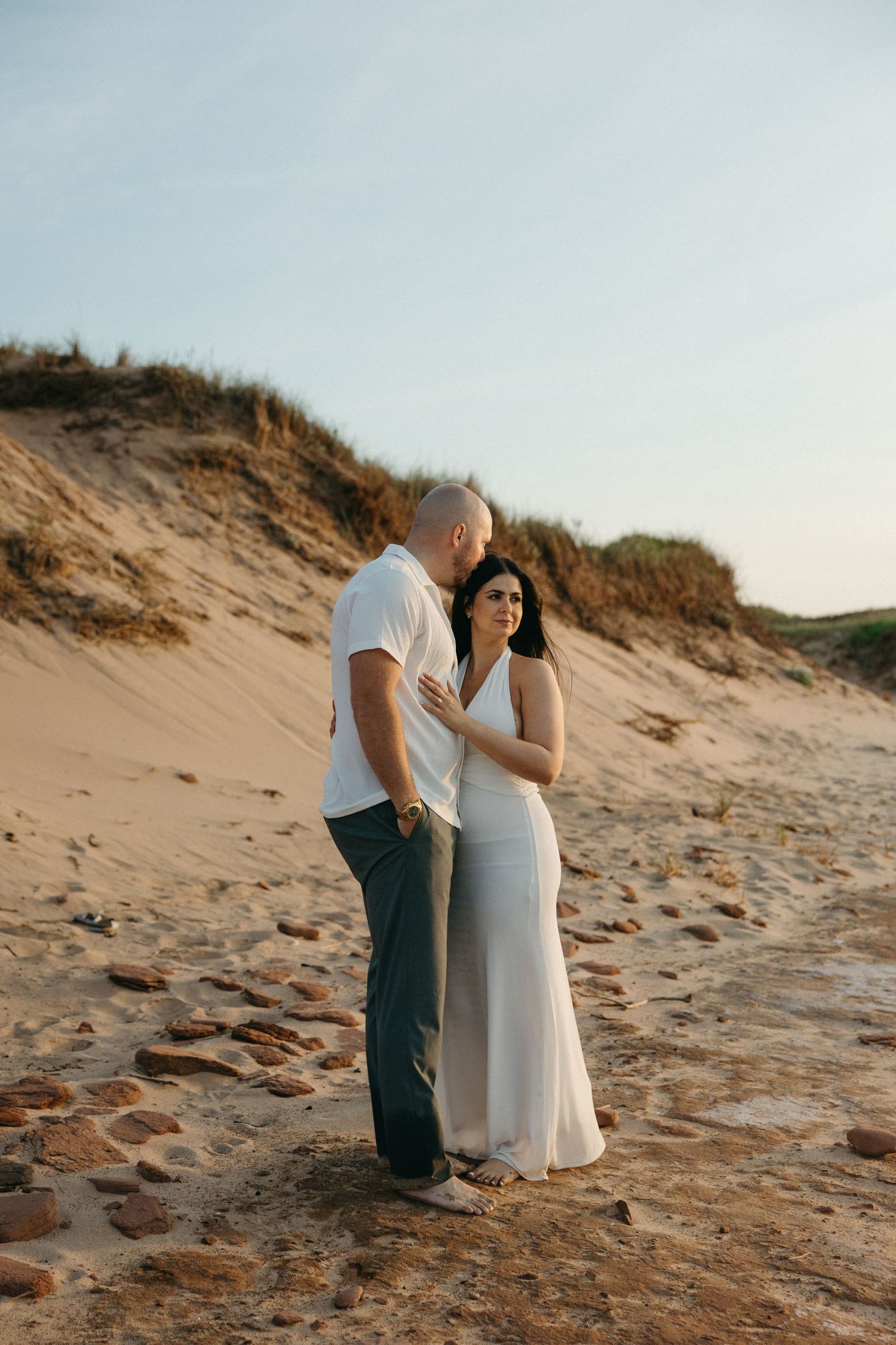 Couple embracing on sandy beach dunes at golden hour, woman in white dress smiling softly.