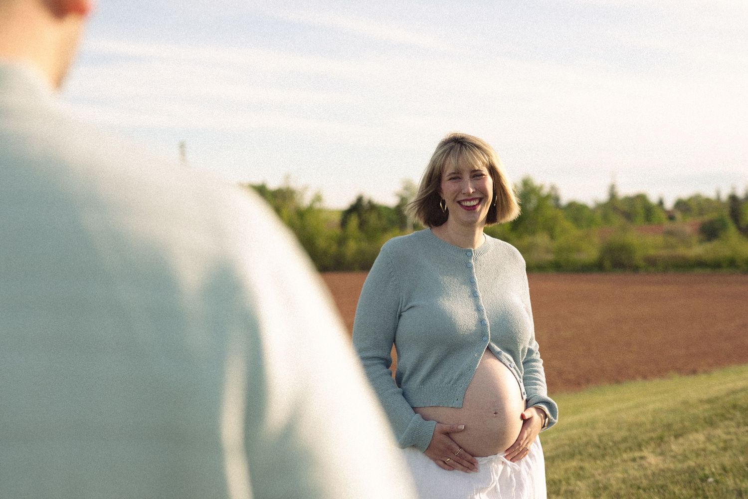 Smiling pregnant woman in light blue cardigan holding her belly in a sunny outdoor field setting.