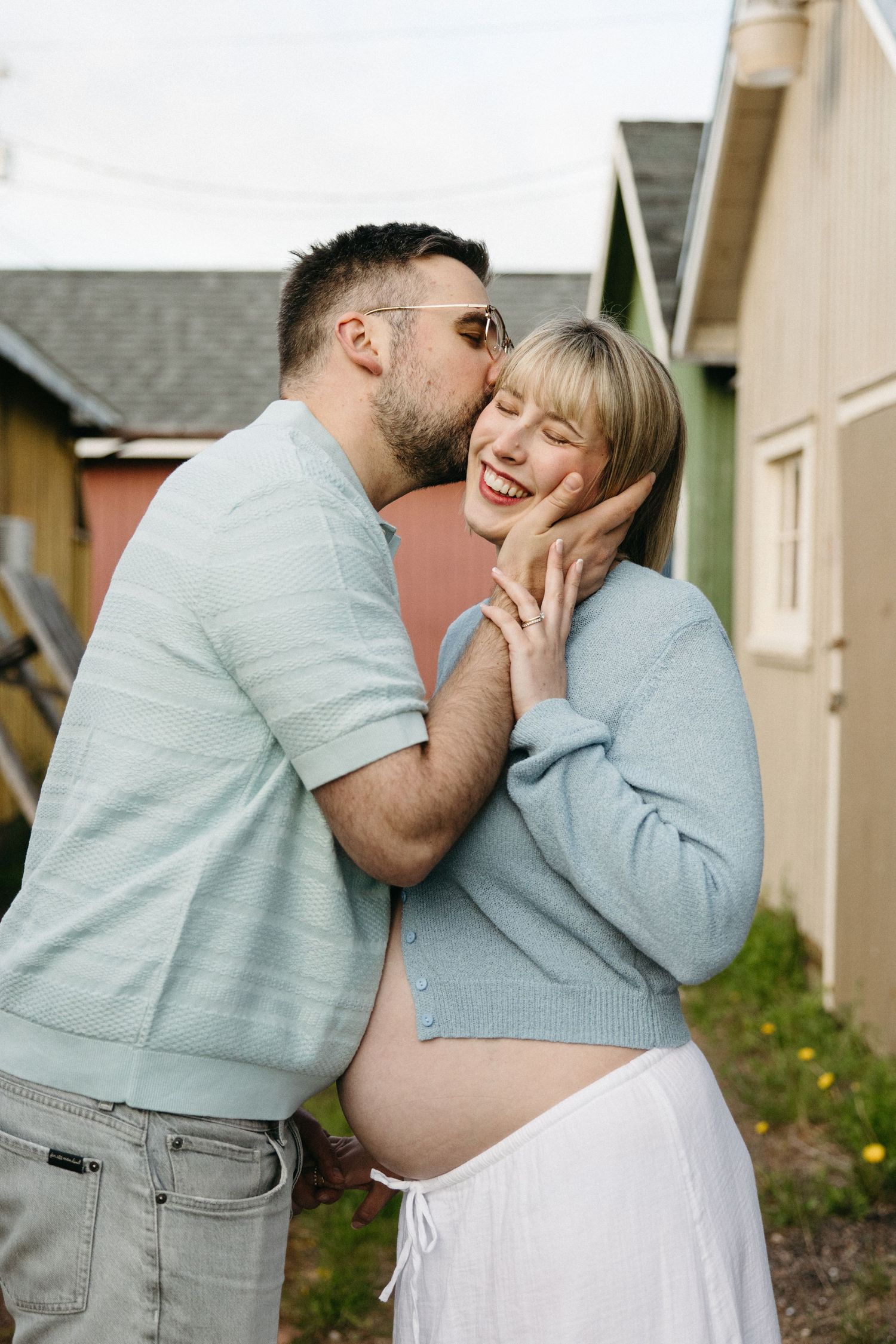 Man kisses laughing pregnant woman on the cheek while she cradles her baby bump near colorful buildings.