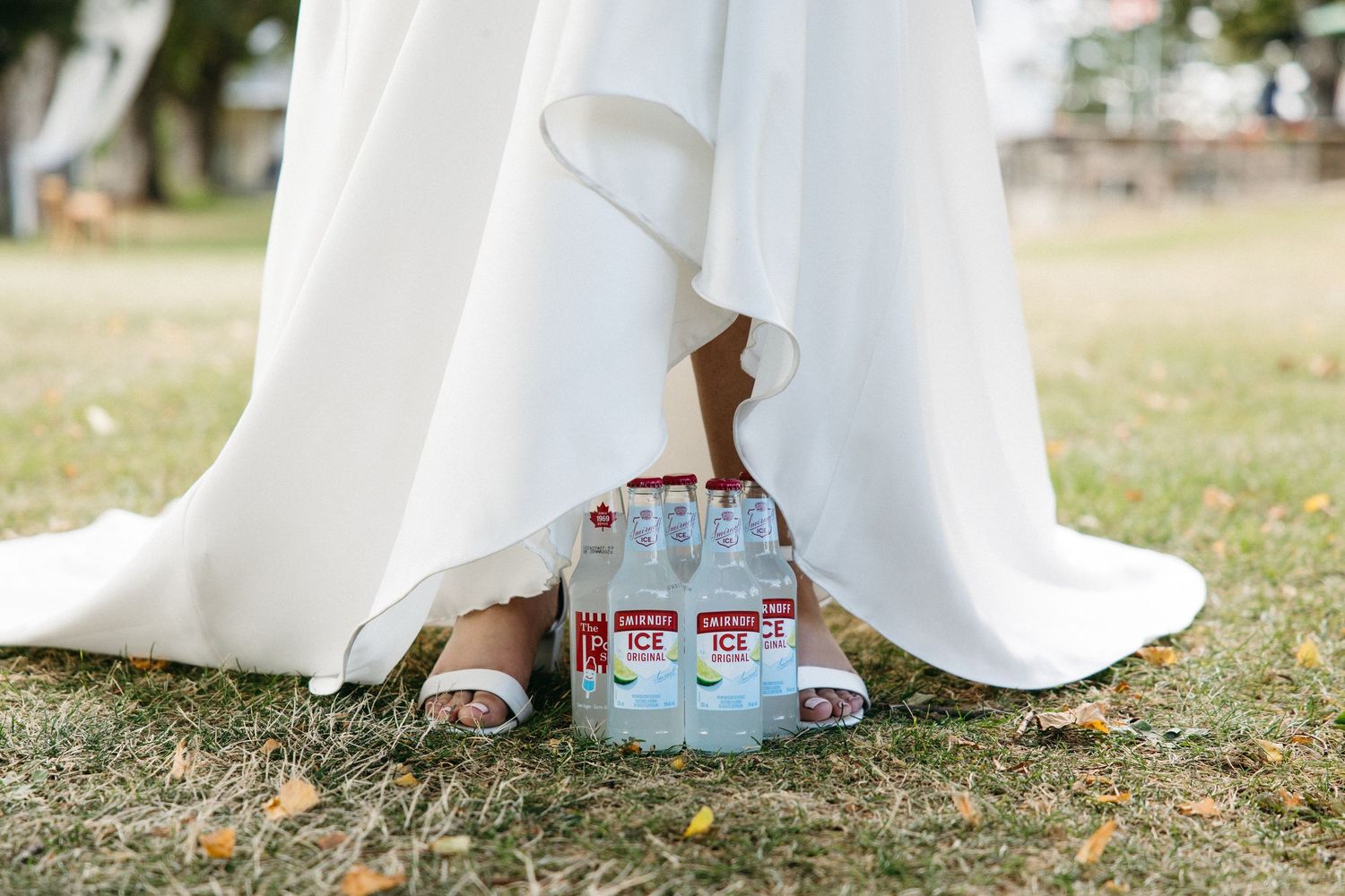 Bride in white wedding gown holds a pack of Smirnoff Ice bottles near her feet on grass.