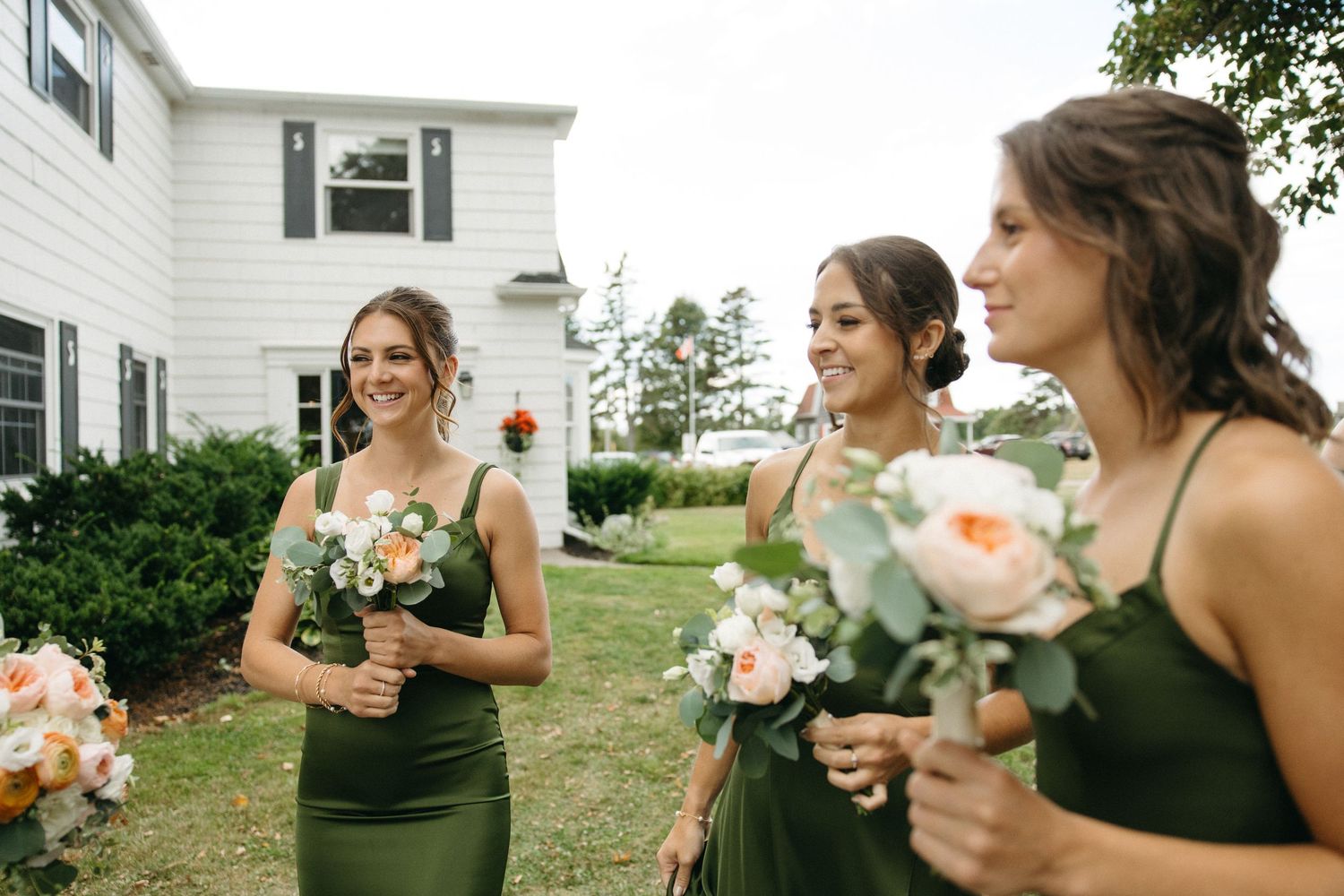Three bridesmaids in olive green dresses holding peach and white bouquets outside a white house.