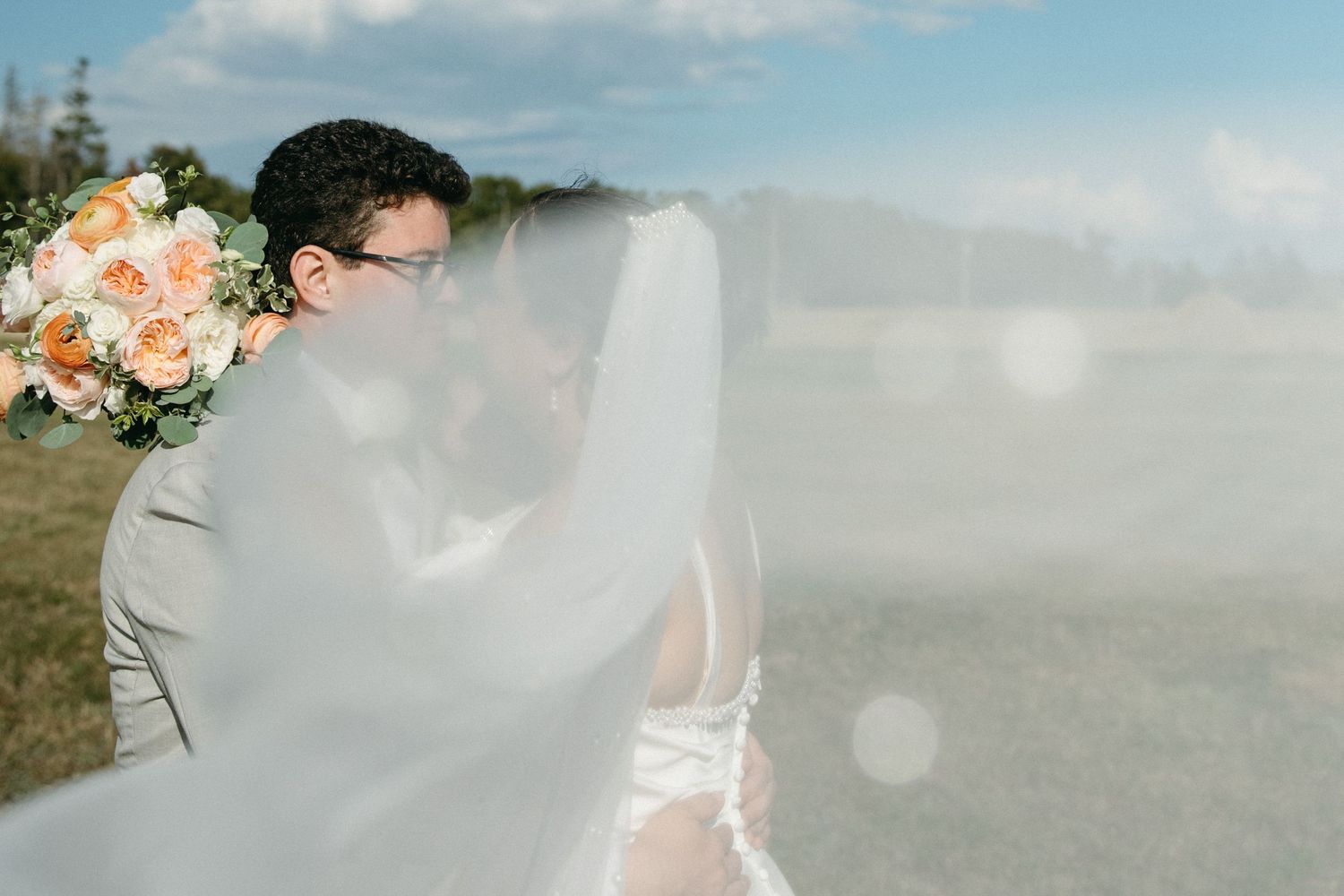 Bride and groom share romantic kiss outdoors with flowing veil and peach bridal bouquet.