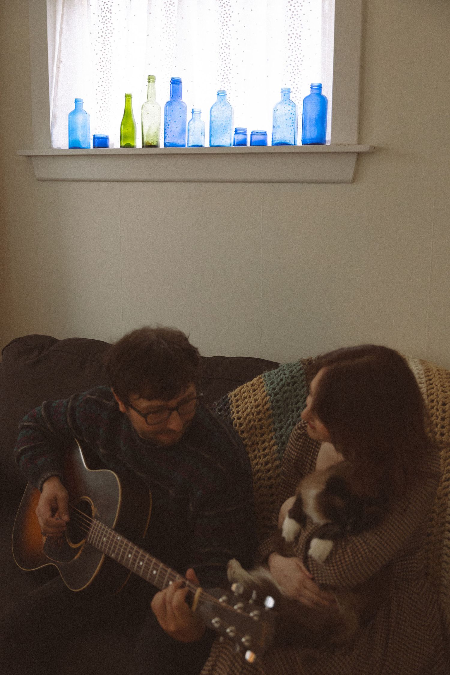 A man playing acoustic guitar on a couch beside a woman holding a cat, with blue glass bottles on a windowsill behind them.
