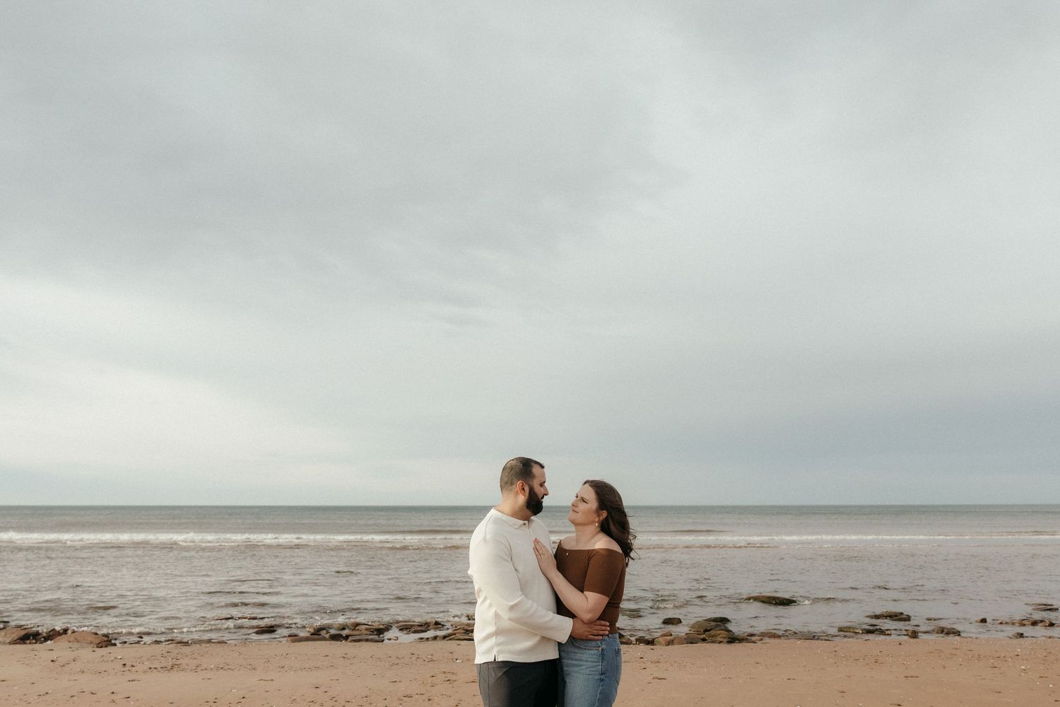 A couple embraces on a rocky beach shoreline during an overcast day.