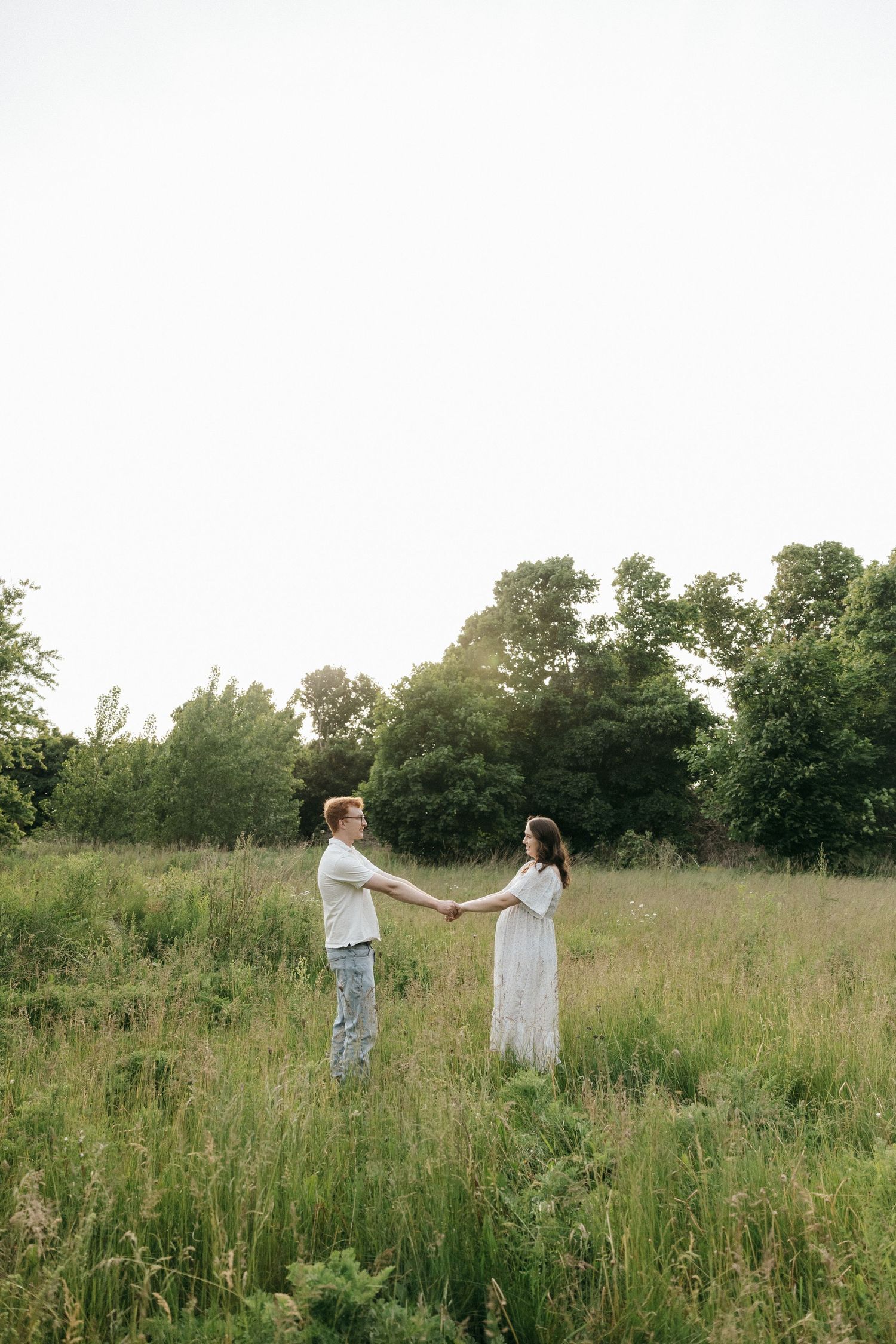 Two figures in light-colored clothing stand together in a sunlit meadow surrounded by tall grass and trees.