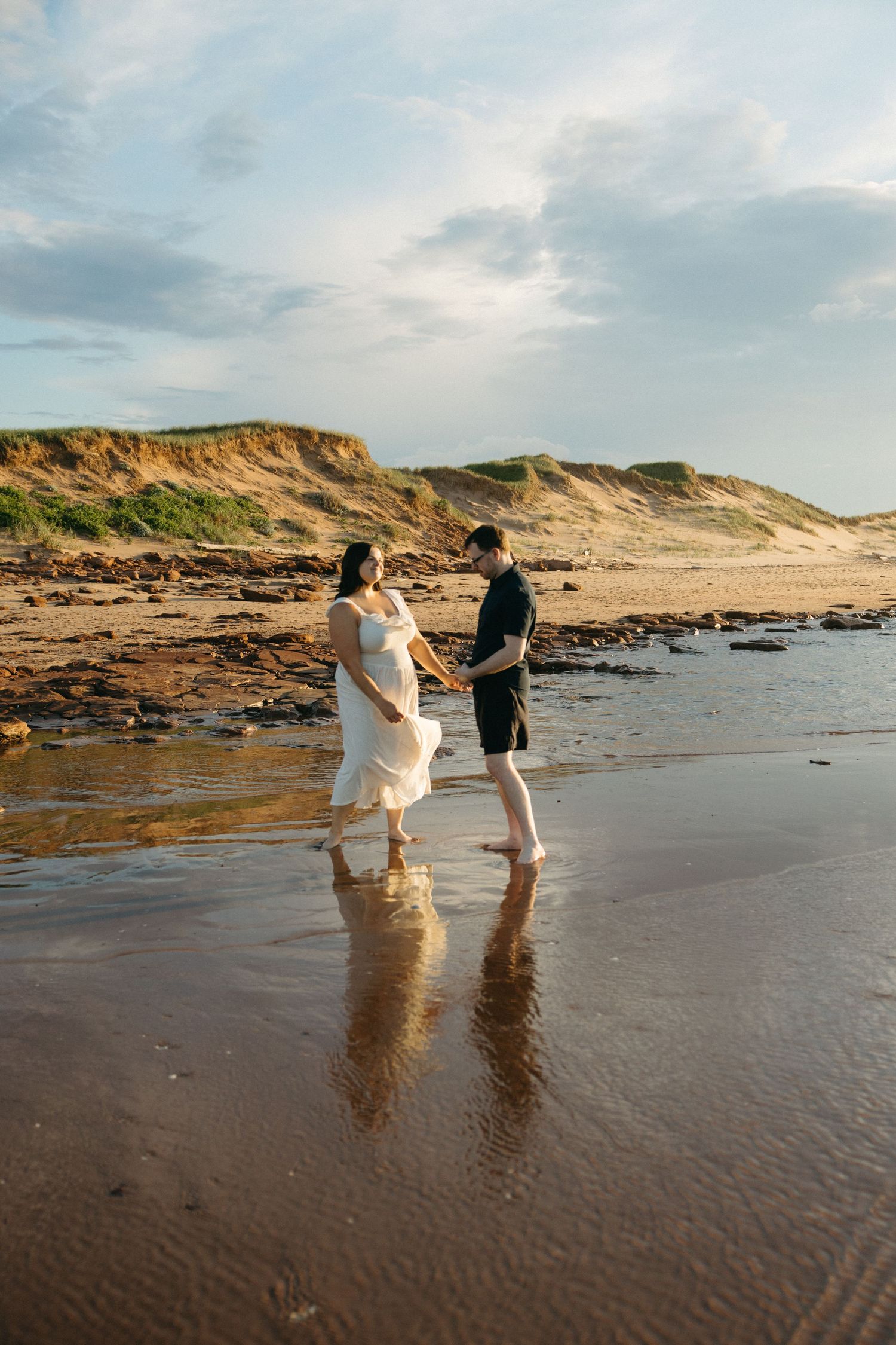 A pregnant couple walks hand in hand along a sandy beach with golden sand dunes in the background.