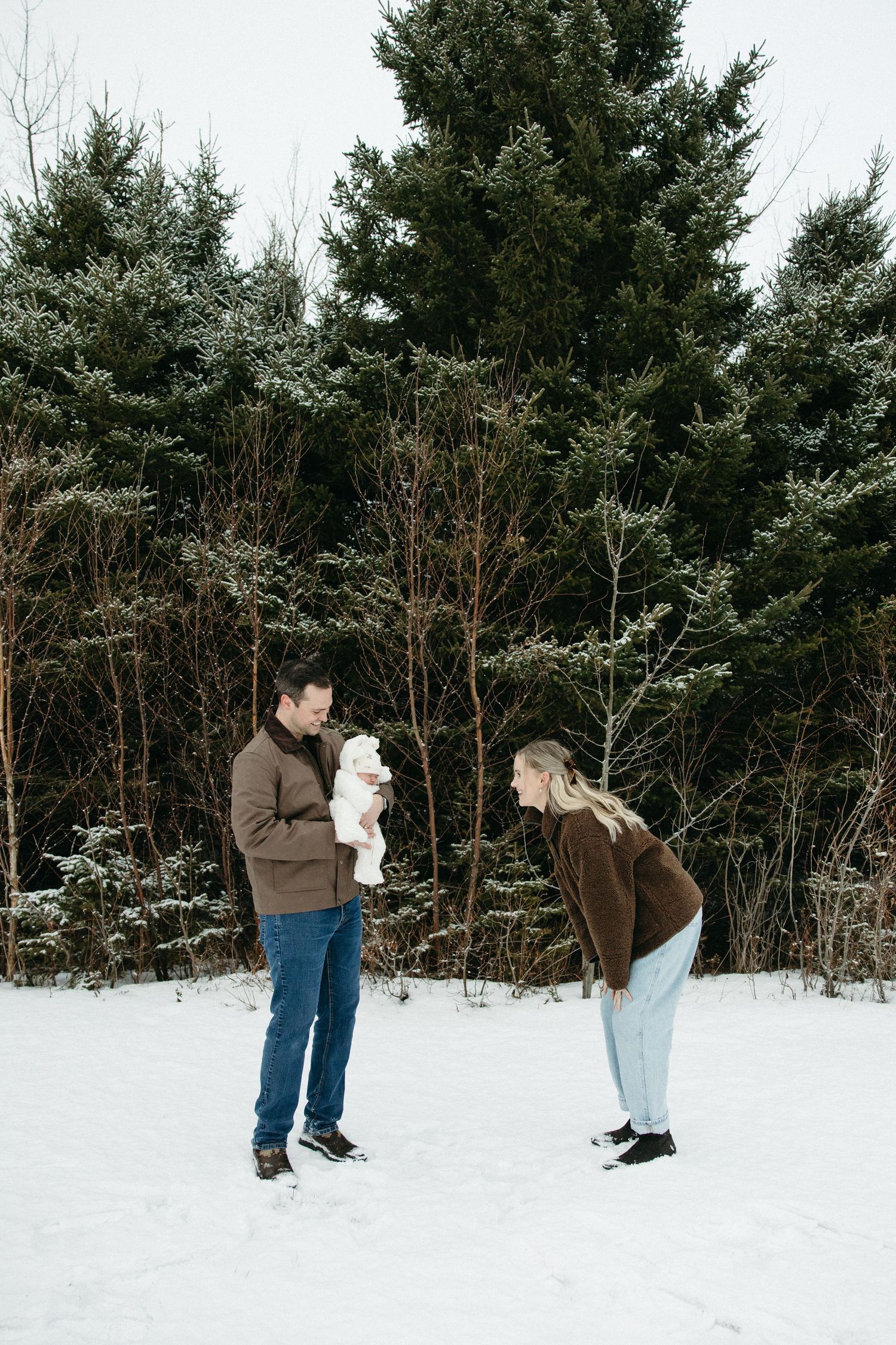A couple interacts playfully with their baby in a snowy forest setting.