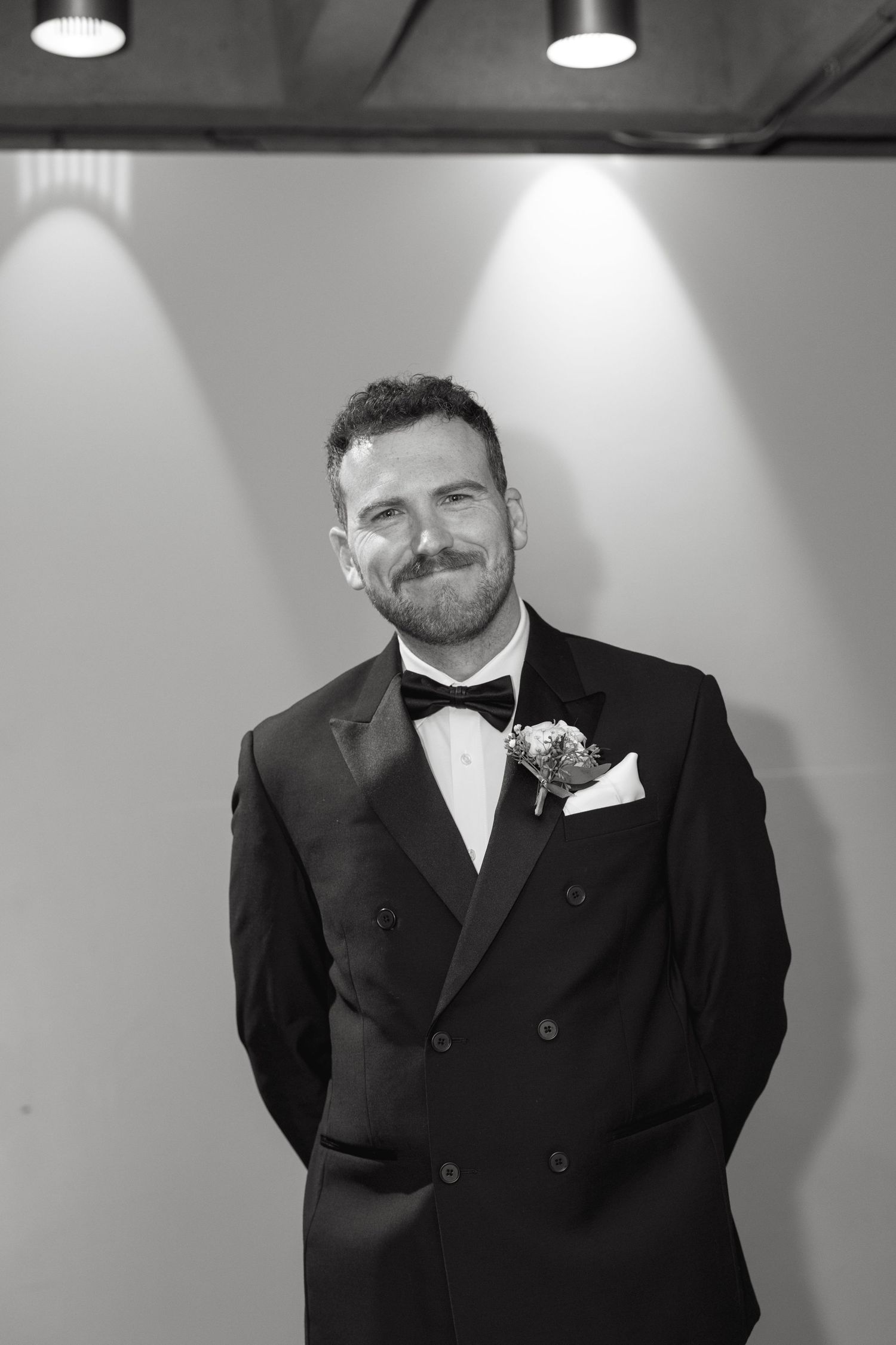 Groom in black tuxedo and bowtie poses against grey wall with dramatic lighting.