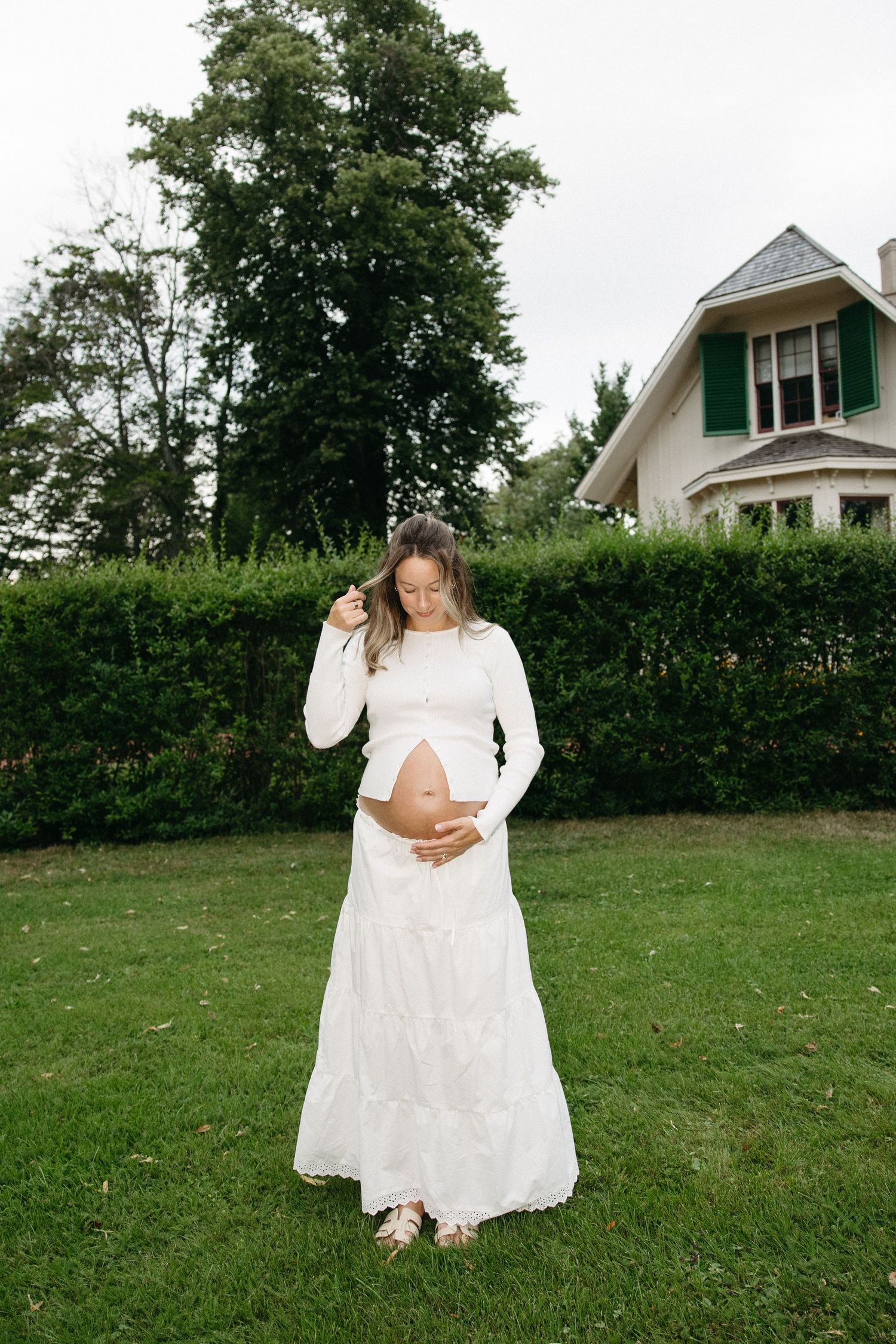 Pregnant woman in white dress cradling baby bump outdoors on green lawn with house and hedges behind her.