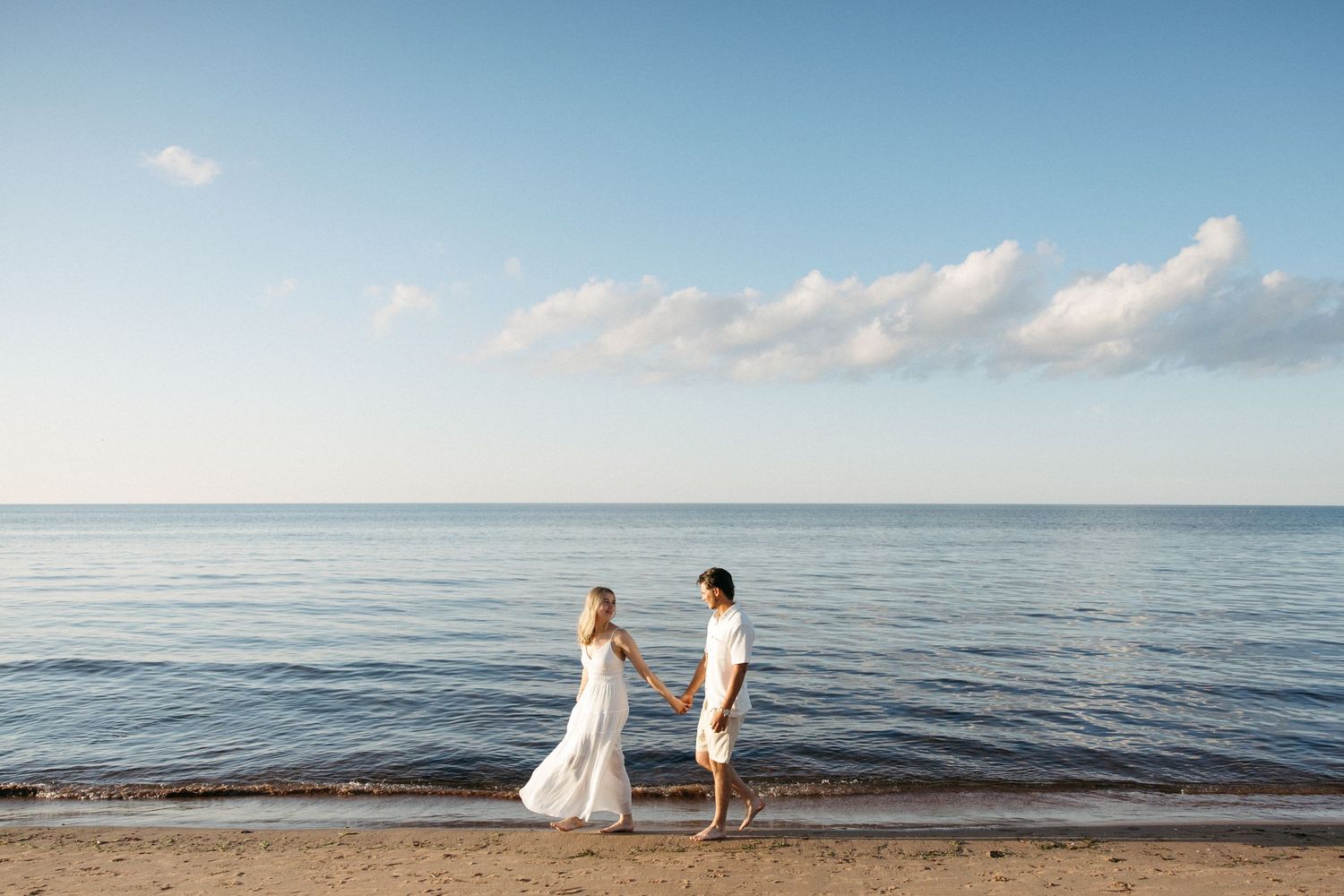 A couple in white holds hands walking along a serene beach with calm blue ocean waters behind them.
