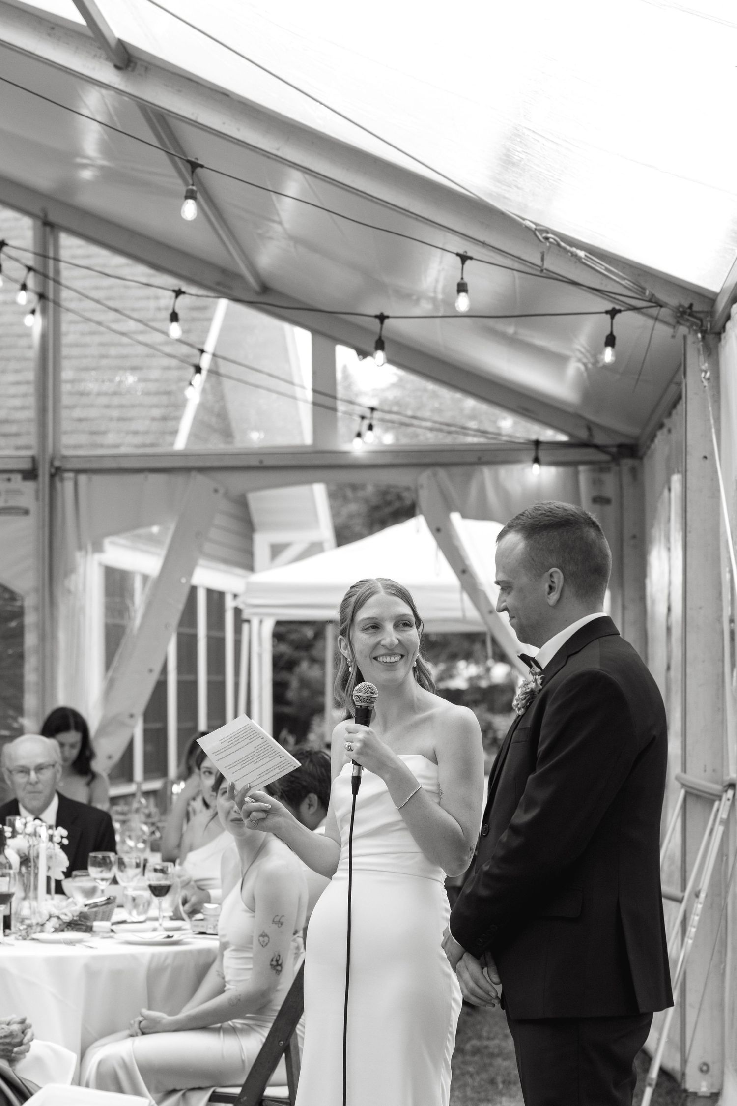 Bride smiles while reading vows to groom under lit wedding tent in black and white photo.