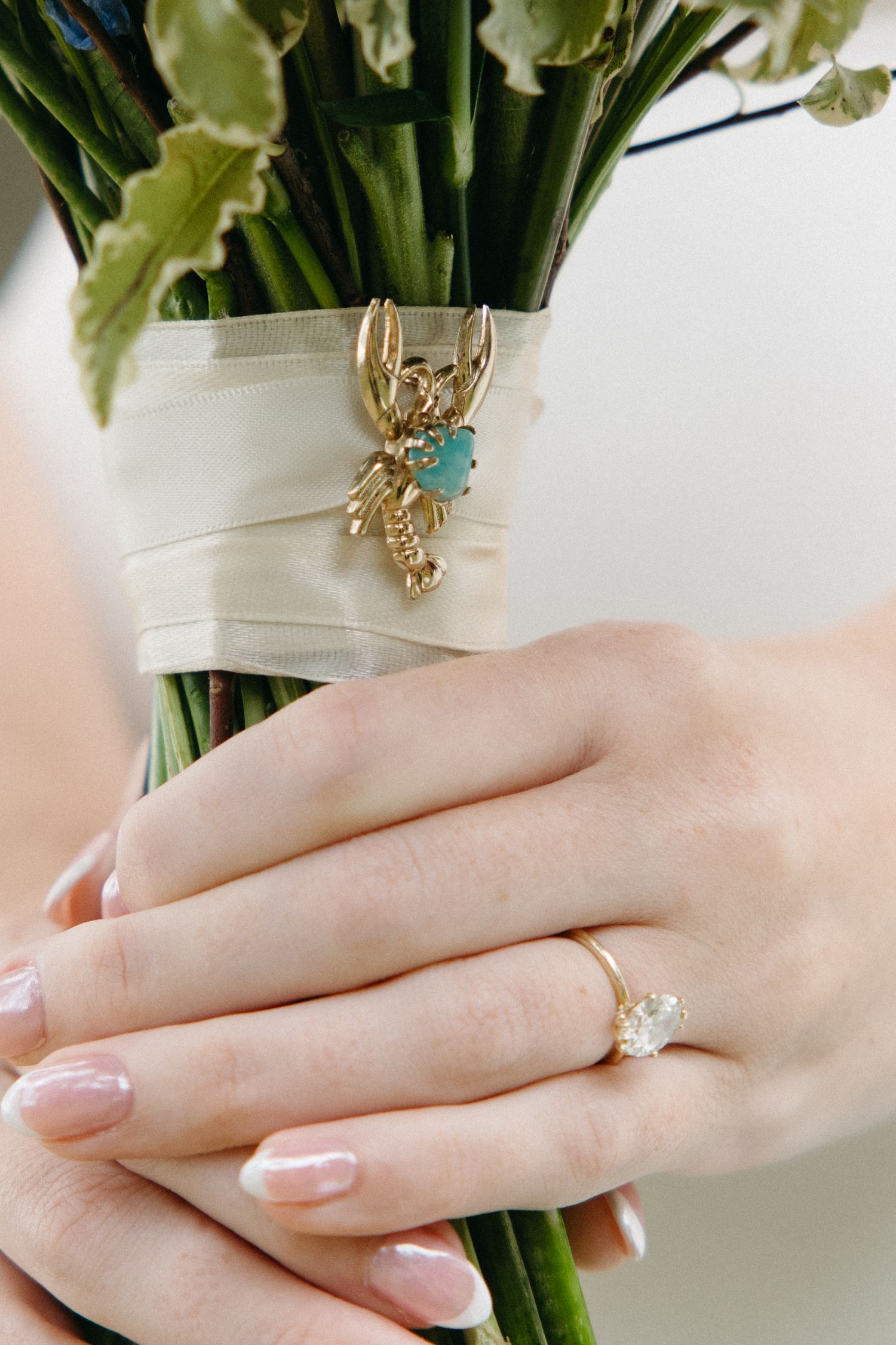 Close-up of bride's hands holding bouquet with gold lobster brooch and diamond engagement ring.
