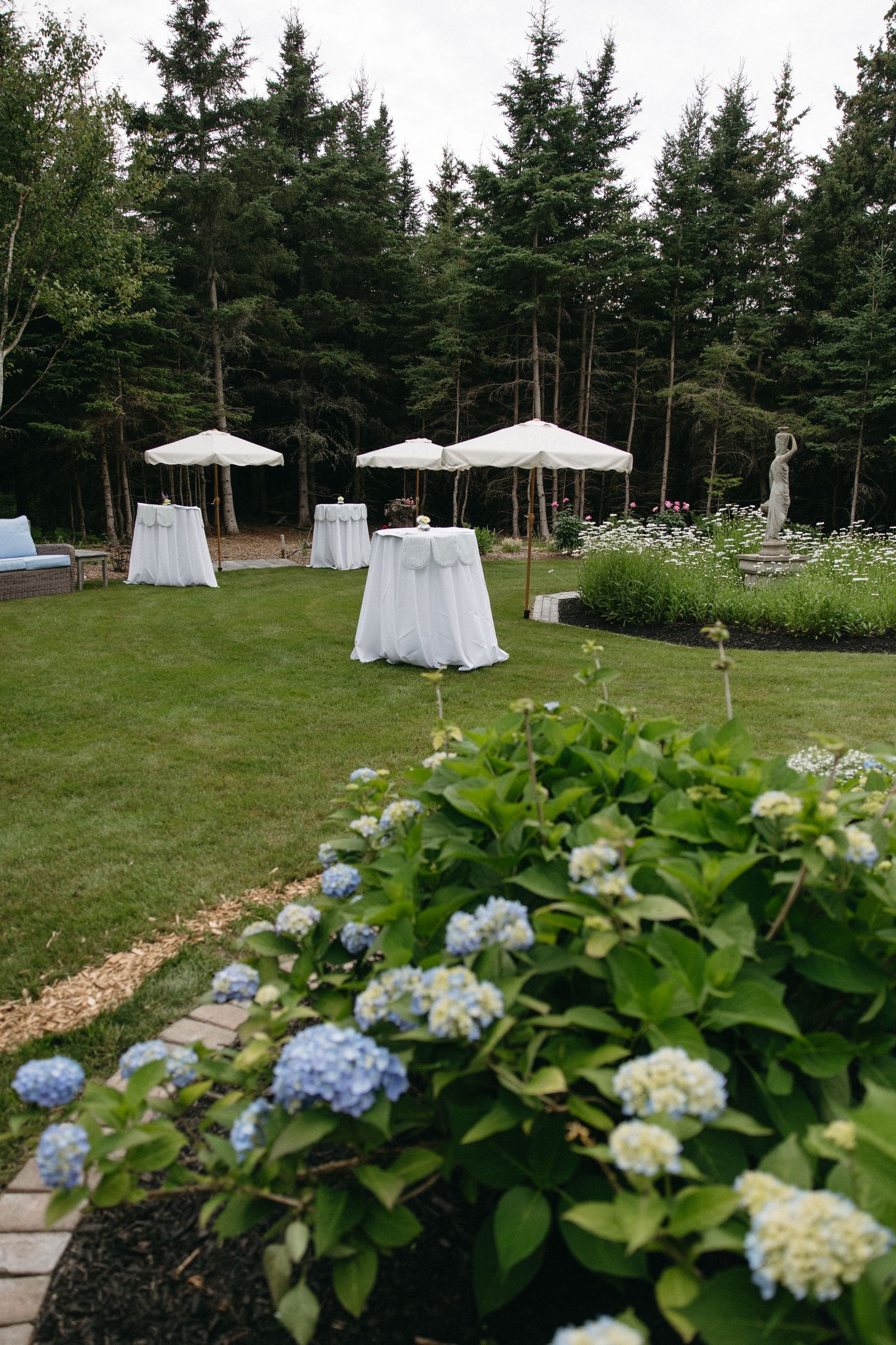 Elegant outdoor wedding cocktail hour setup with white draped tables, umbrellas, and blue hydrangeas in foreground.