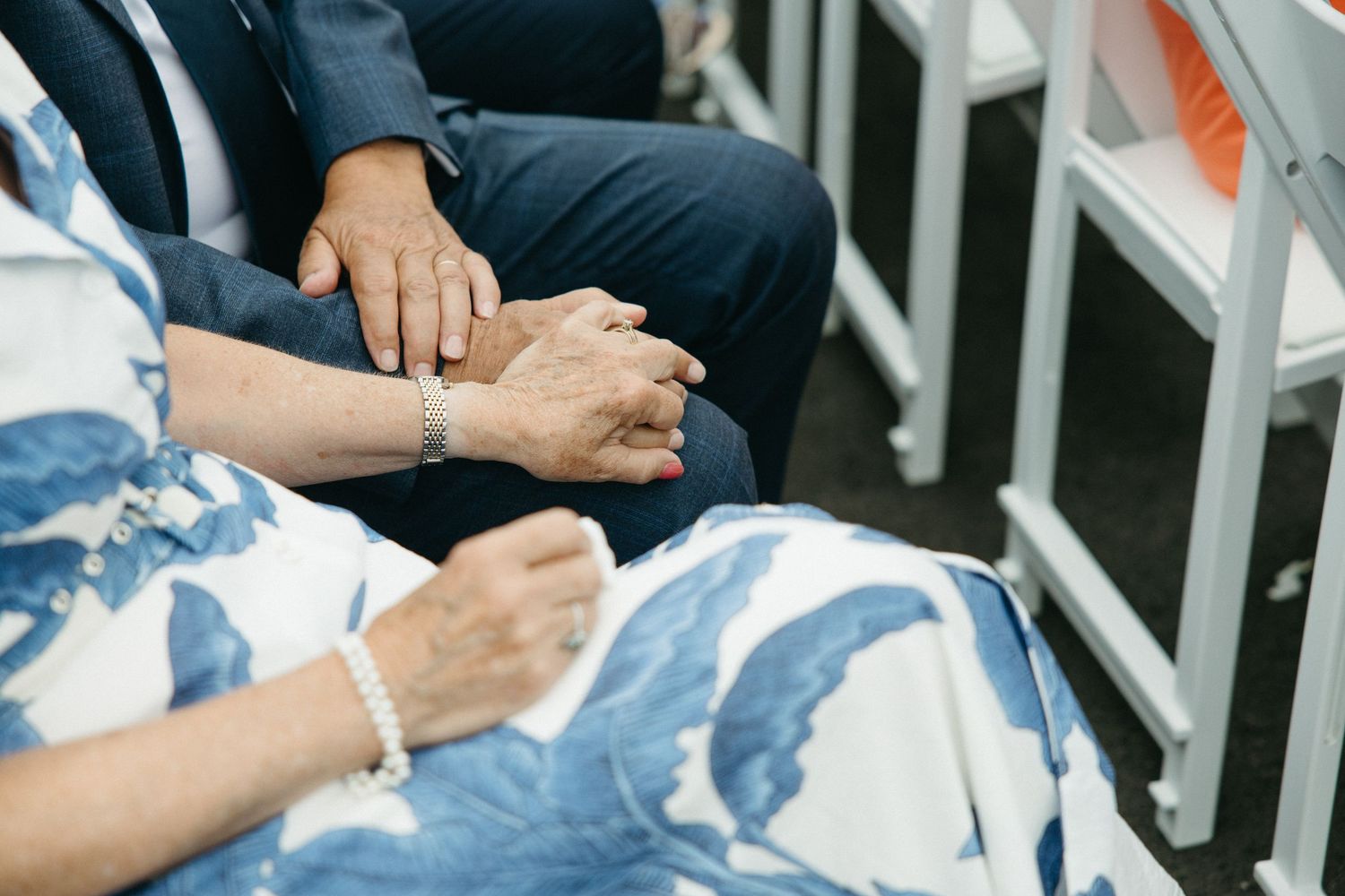 Elderly couple holding hands tenderly while seated at an outdoor wedding ceremony.