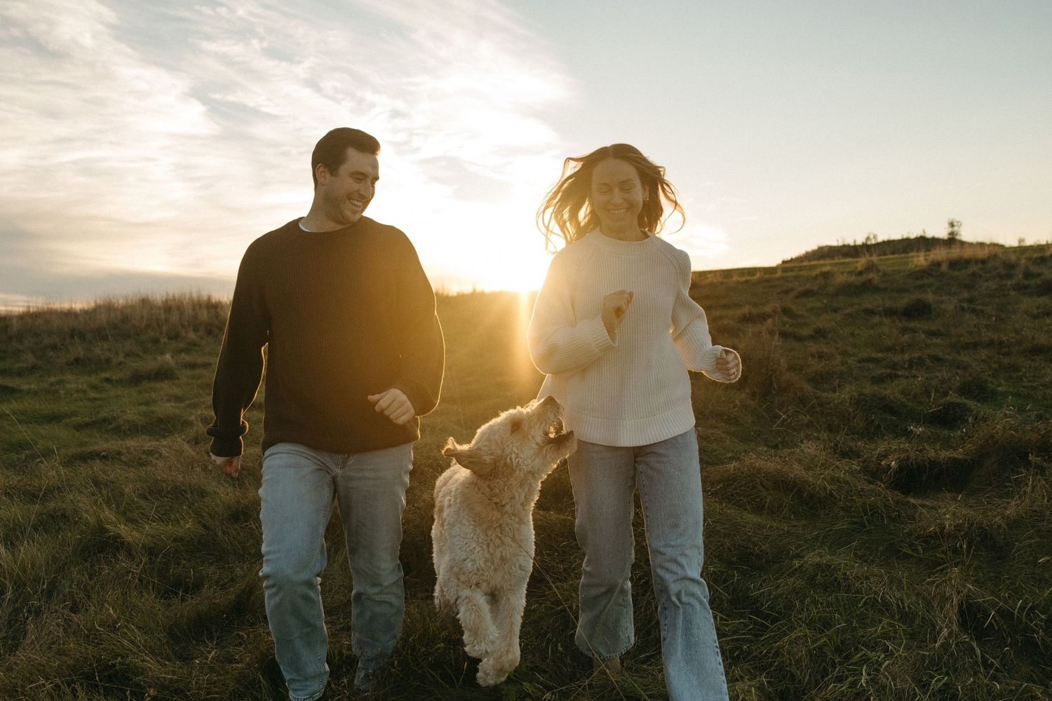 Smiling couple walking with a fluffy cream-colored dog across a grassy hillside at golden hour sunset.