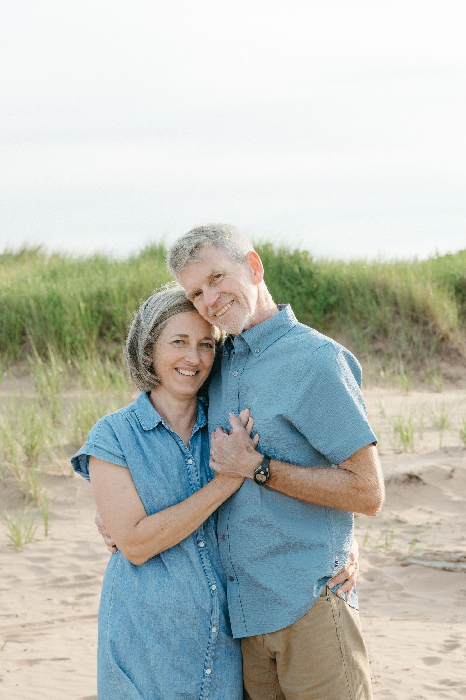 A happy middle-aged couple embraces and smiles on a sandy beach with green dune grass behind them.