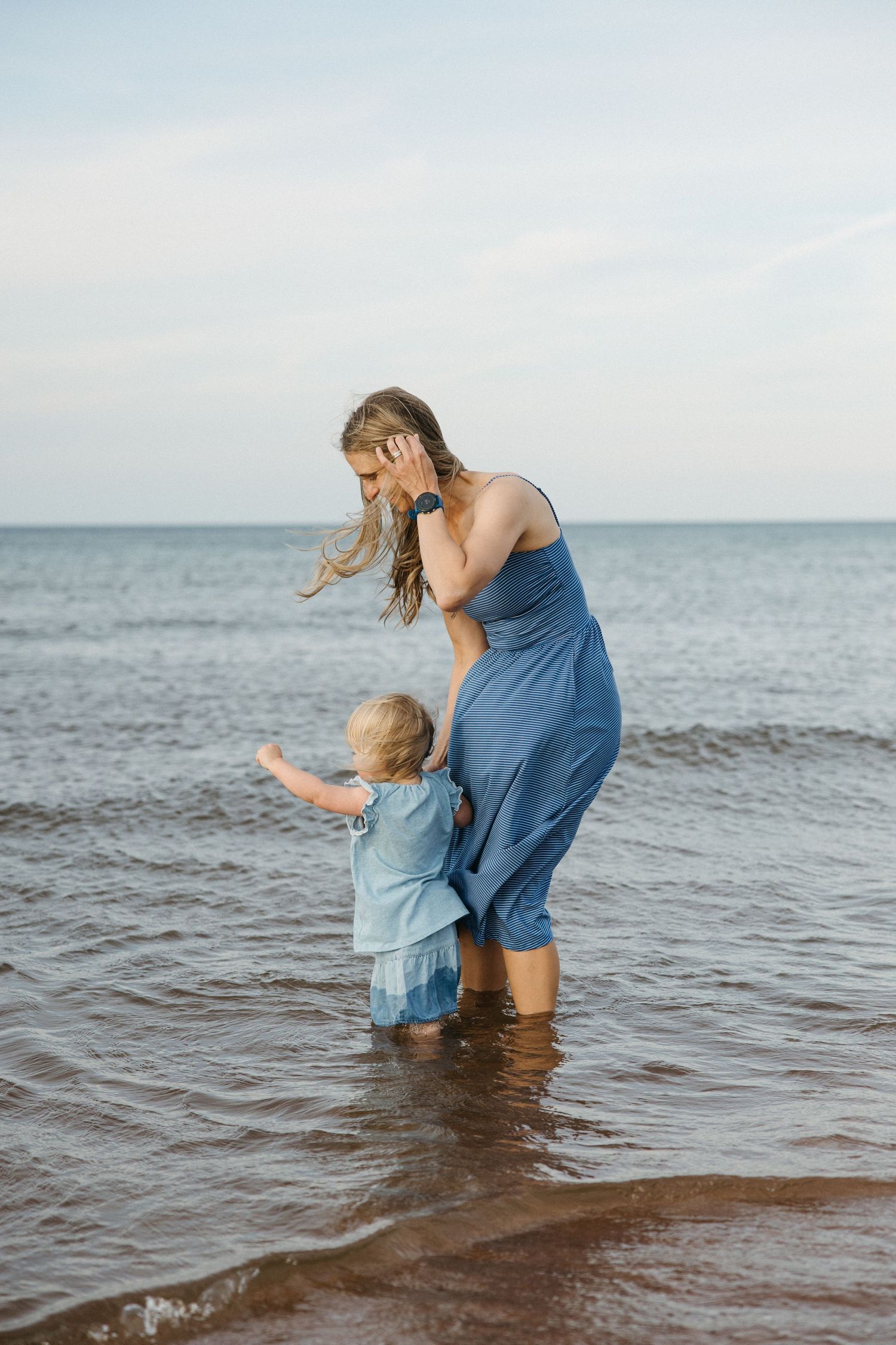 Mother in blue dress helping toddler walk through shallow ocean waves at beach.