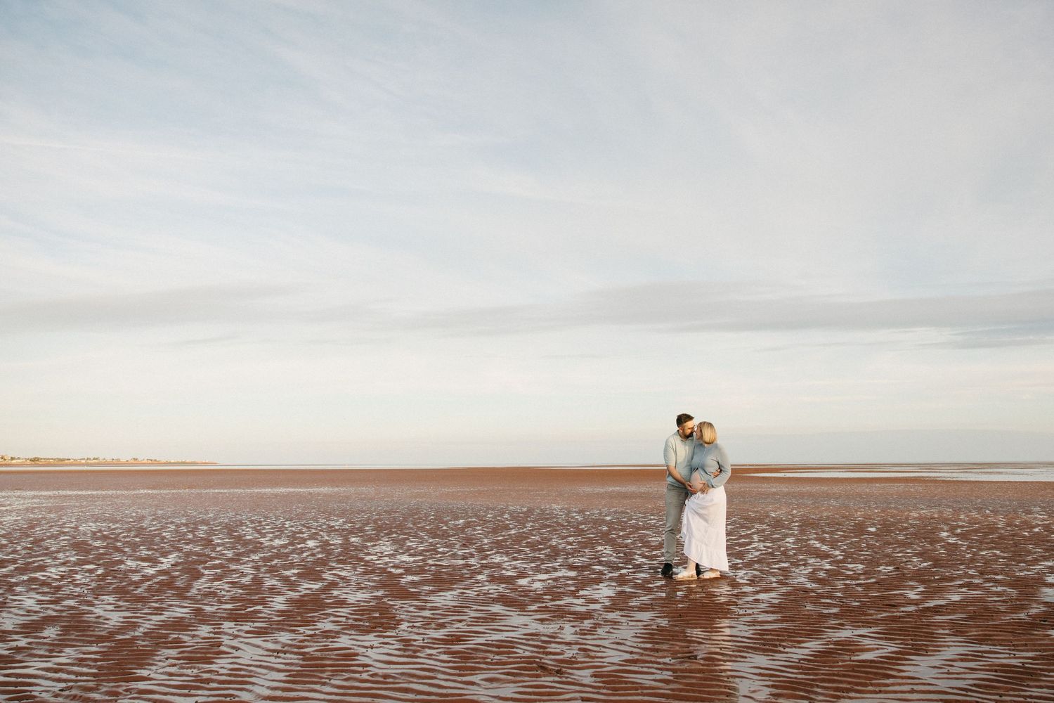 Couple embracing on a vast, reflective beach at golden hour with a wide open sky backdrop.