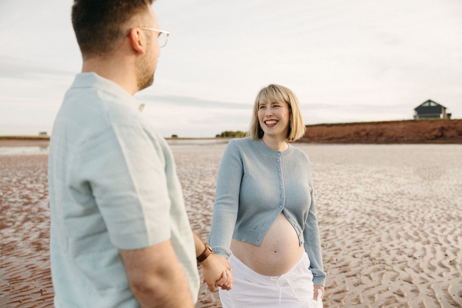 Pregnant woman laughing with partner on sandy beach, holding hands during maternity photoshoot at sunset.