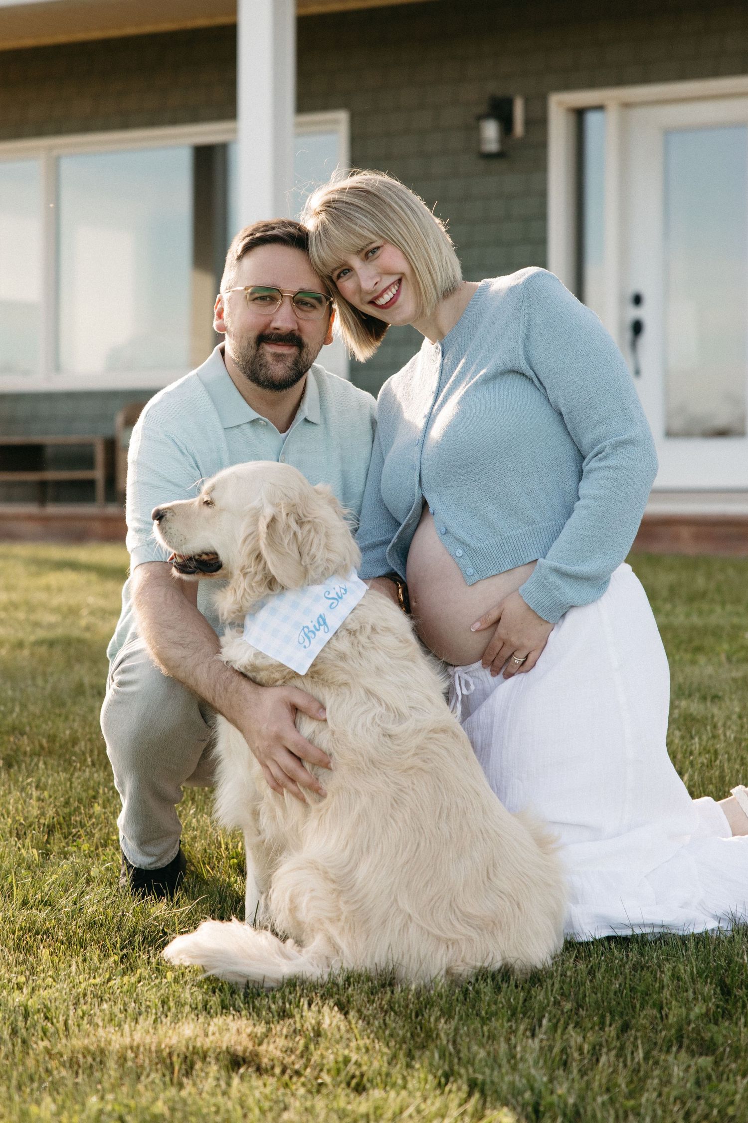 Smiling pregnant couple poses outdoors with their golden retriever wearing a blue bandana, sitting on grass in front of a house.