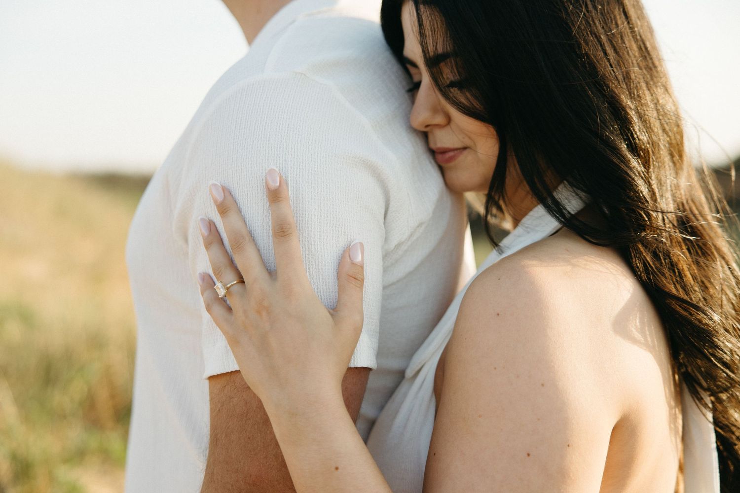 A woman with dark hair embraces a man in a white shirt outdoors, showcasing her diamond engagement ring.