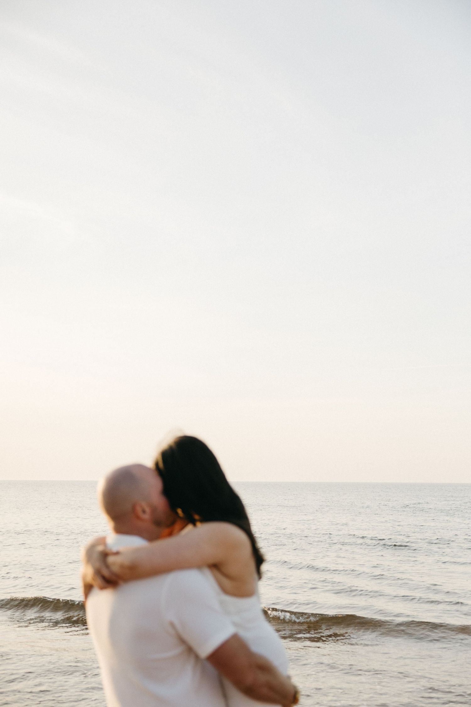 Couple embracing and kissing on the beach with serene ocean and soft sky in the background.