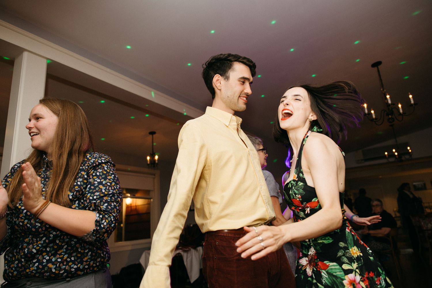 Guests laugh and dance together under colorful lights at lively wedding reception.