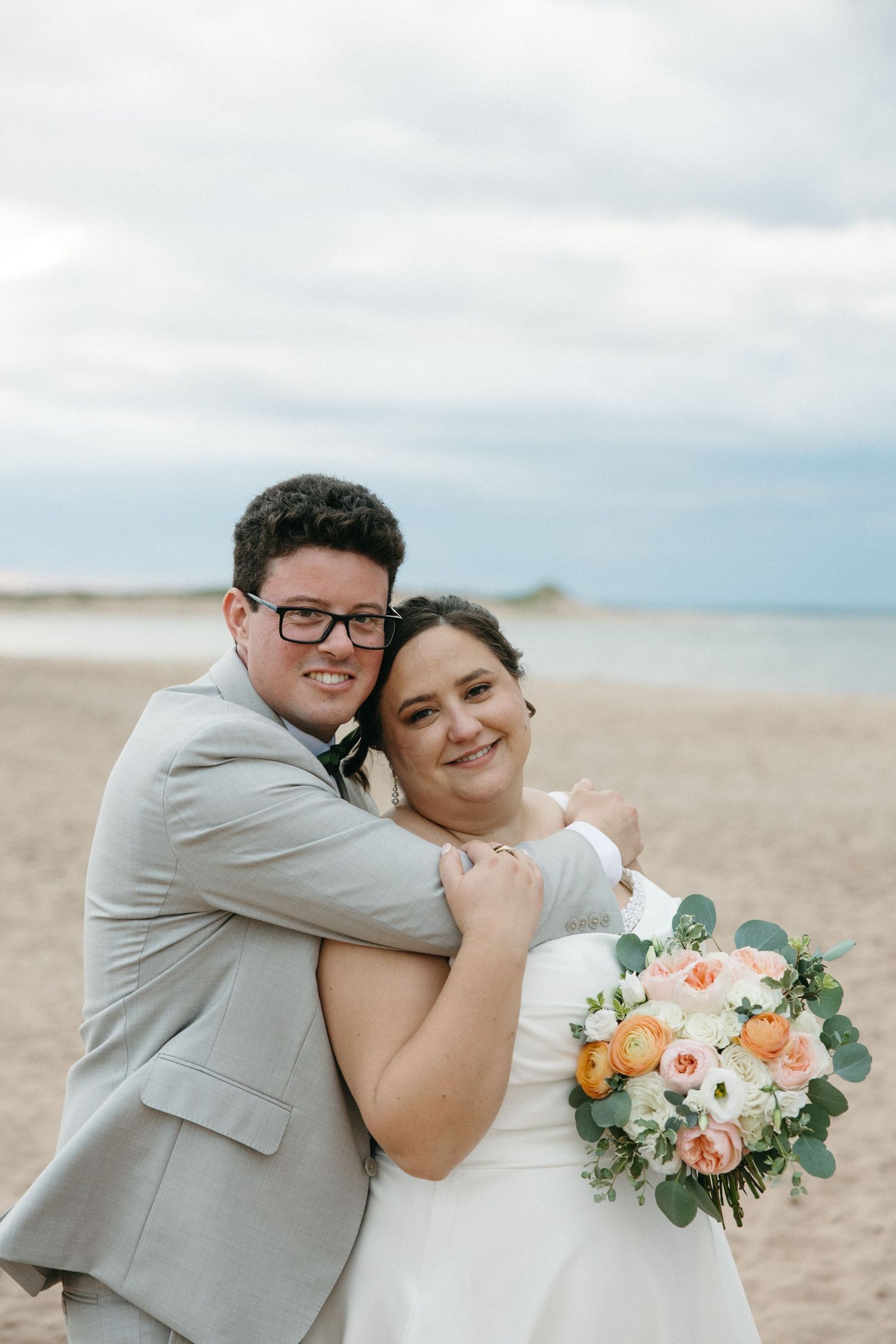 Happy newlywed couple embraces on a sandy beach, groom in gray suit holding bride with peach bouquet.