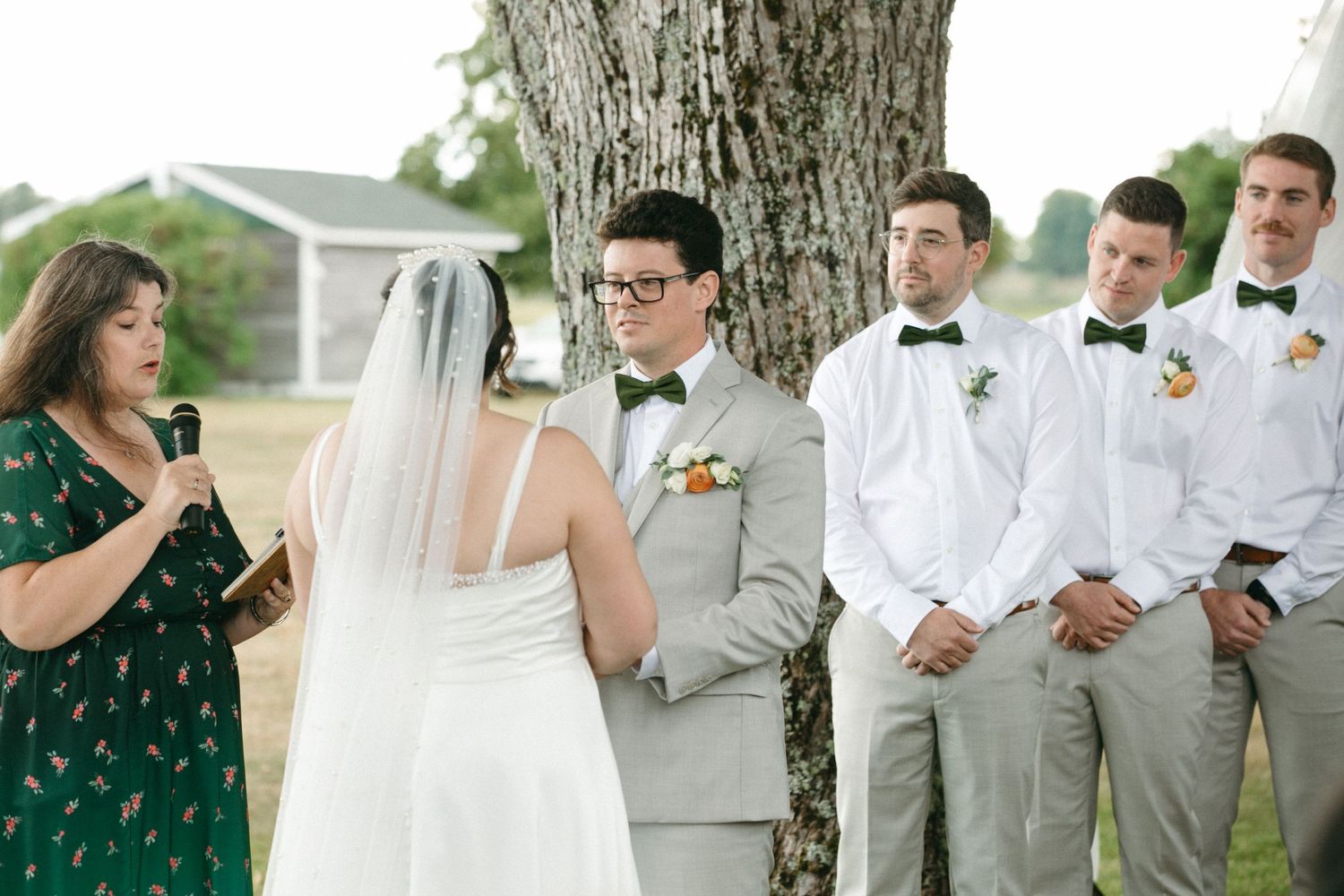 Bride and groom exchanging vows under a large tree with groomsmen in bow ties standing nearby.