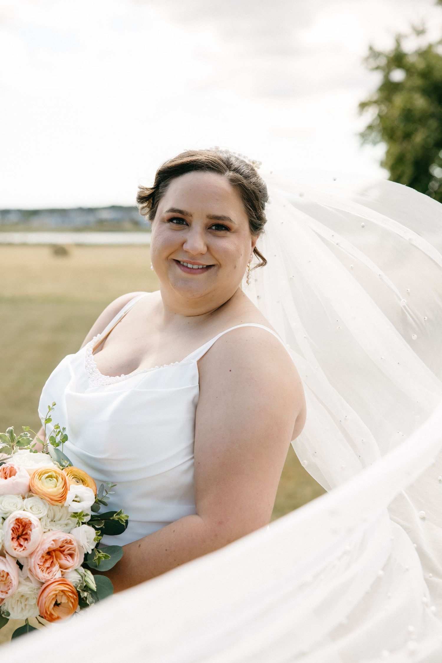 Smiling bride in white spaghetti strap gown holds peach and white floral bouquet with veil flowing in wind.