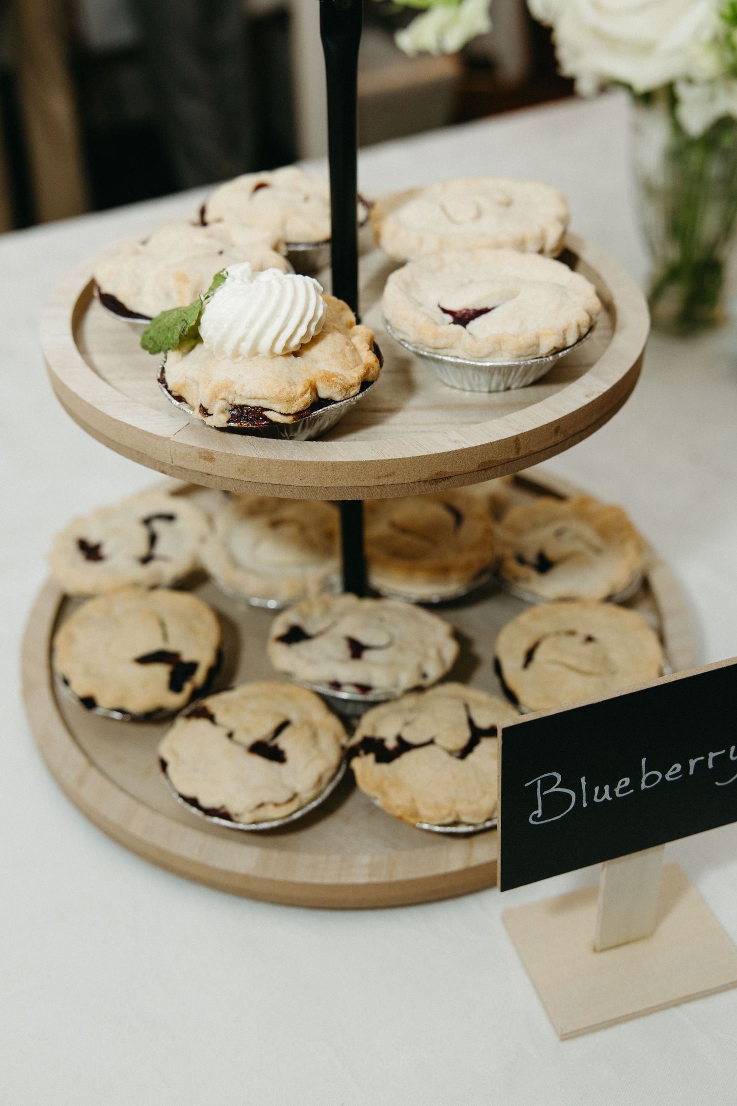 Two-tiered wooden dessert stand displaying mini blueberry pies at a wedding reception.