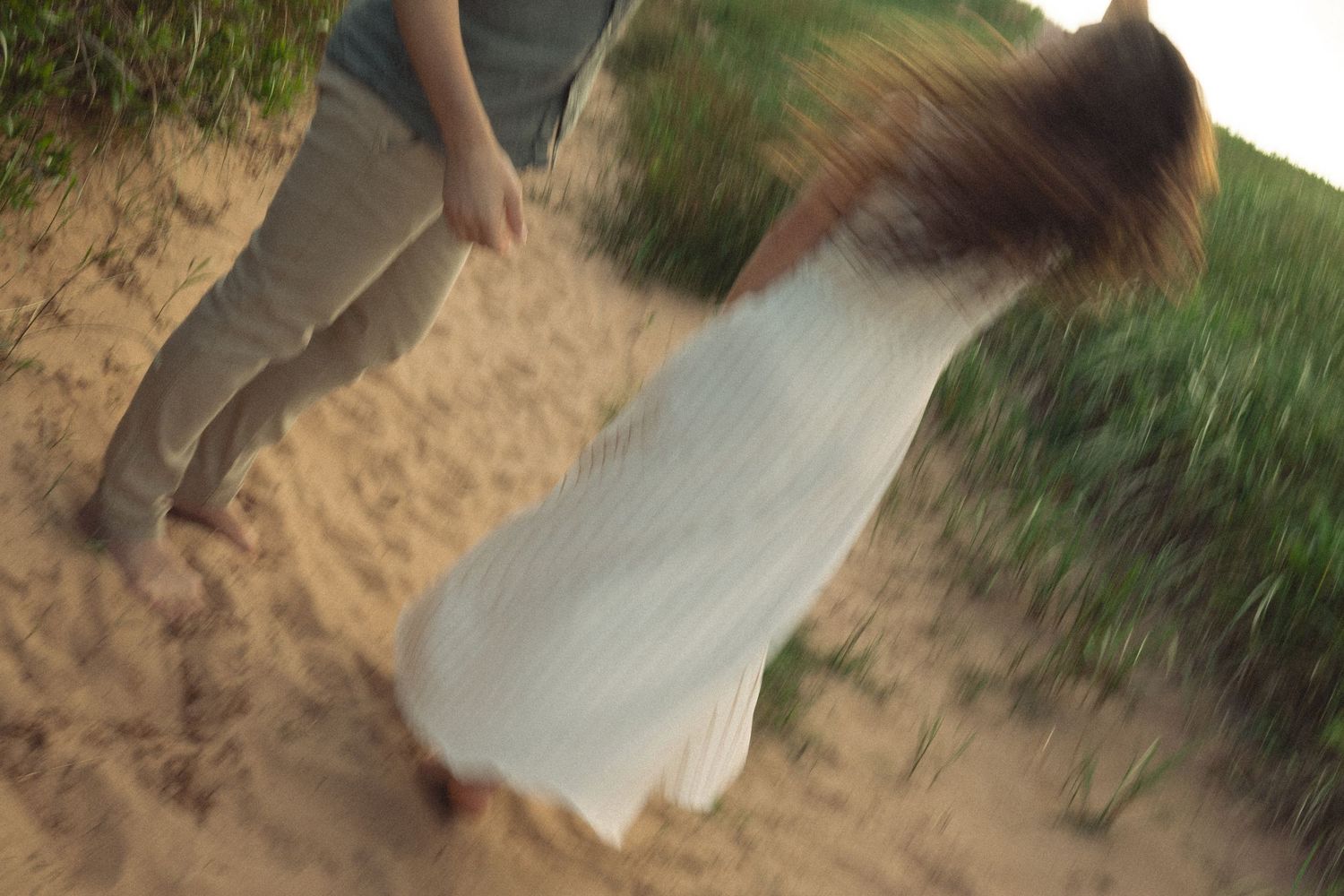 Couple walking barefoot on sandy beach dunes, woman in white dress with flowing hair, motion blur effect.