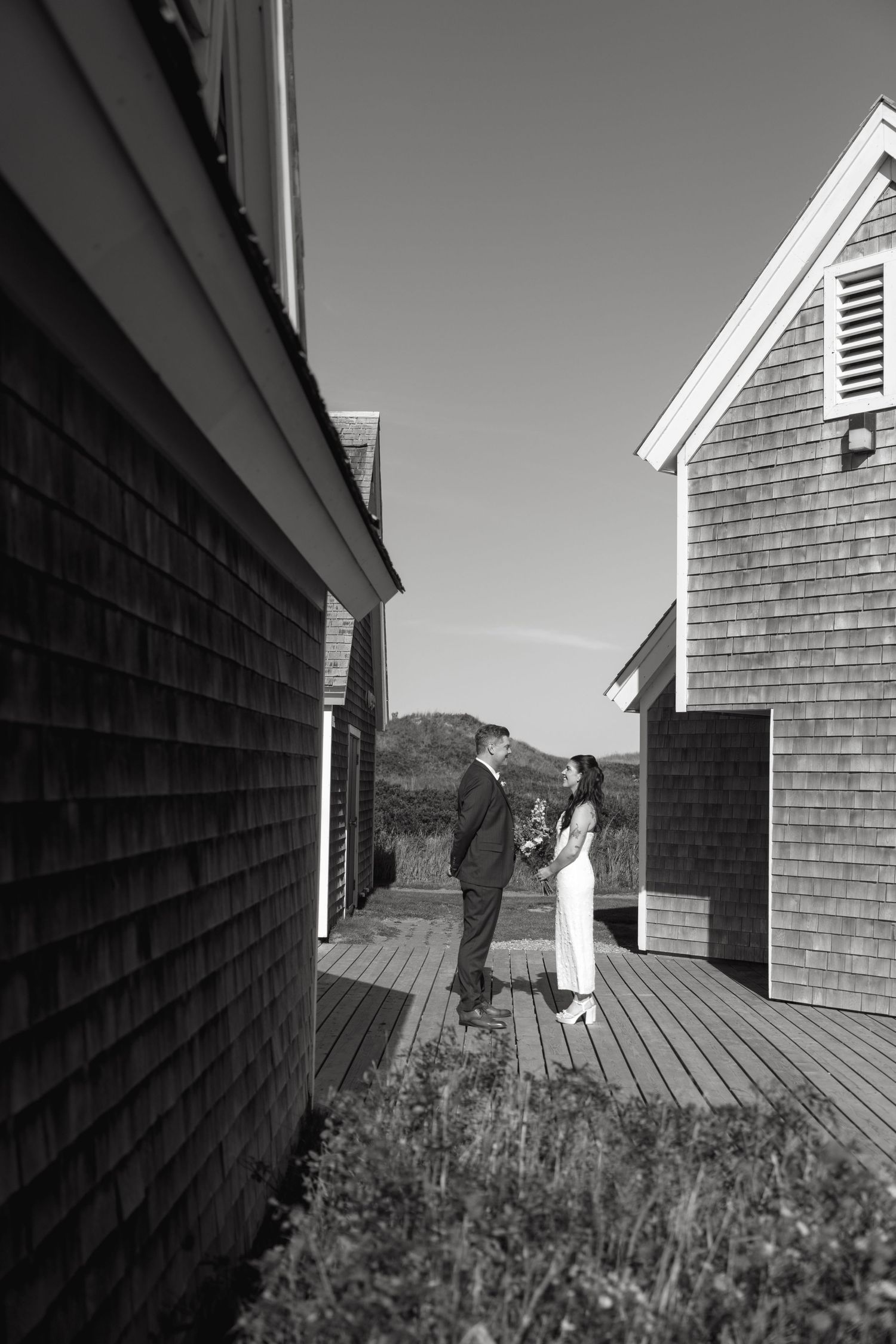 Black and white photo of a bride and groom facing each other on a wooden walkway between weathered cedar-shingled cottages.