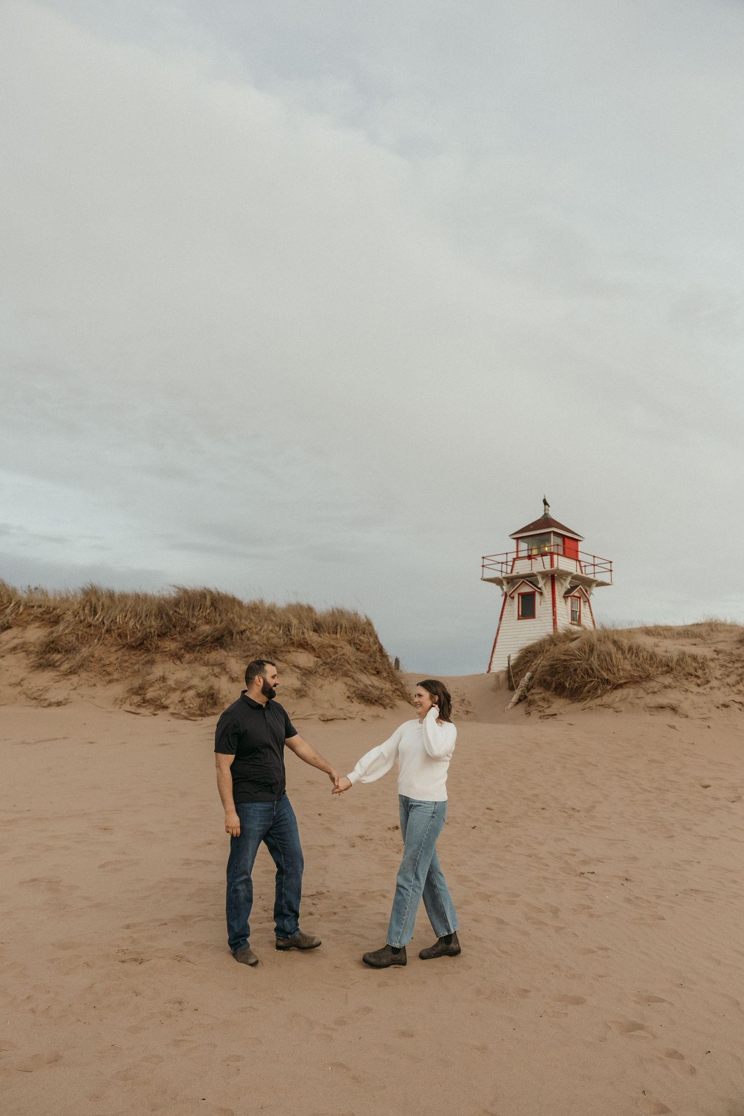 Two people walk together on sandy dunes with a lighthouse tower visible in the distance on an overcast day.