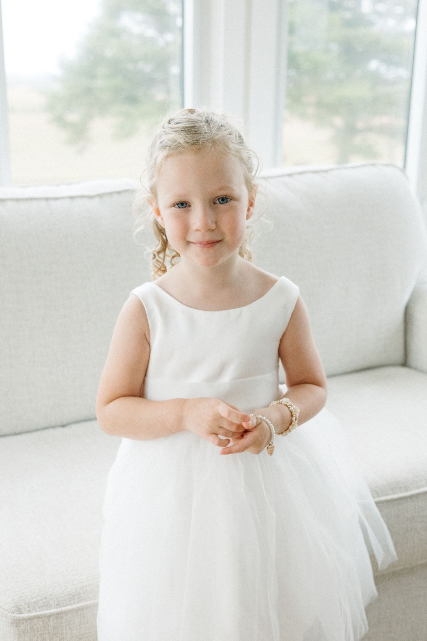 Young flower girl in white dress sitting on light colored couch.