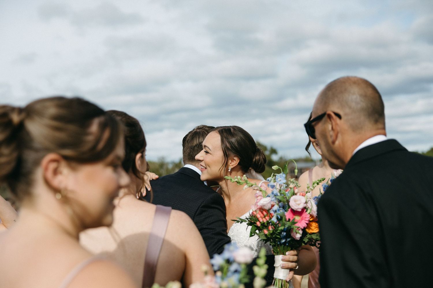 Wedding party gathered outdoors with colorful bouquets against a cloudy sky.