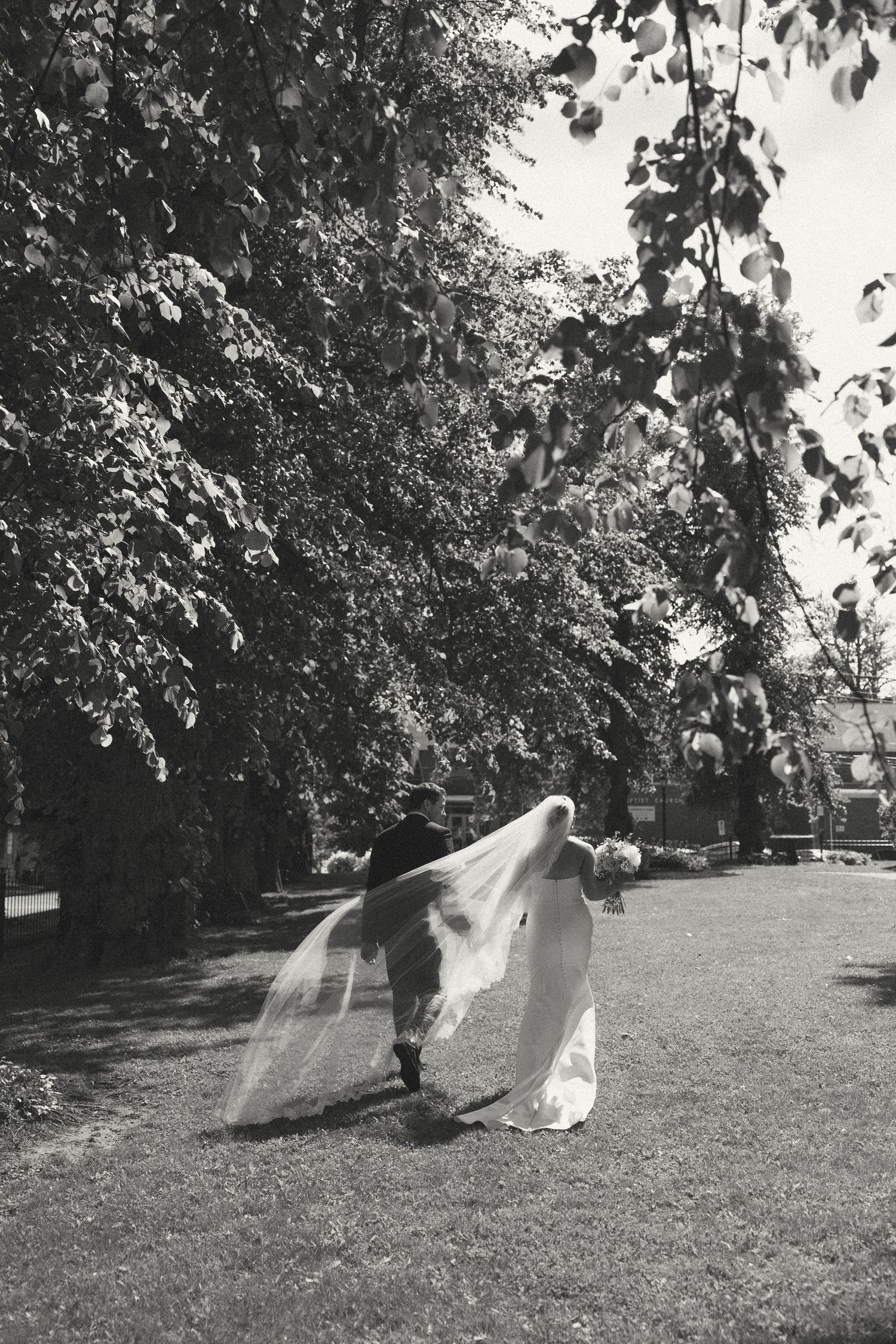 Artistic black and white photo of a wedding dress veil flowing in the breeze on a grassy lawn.