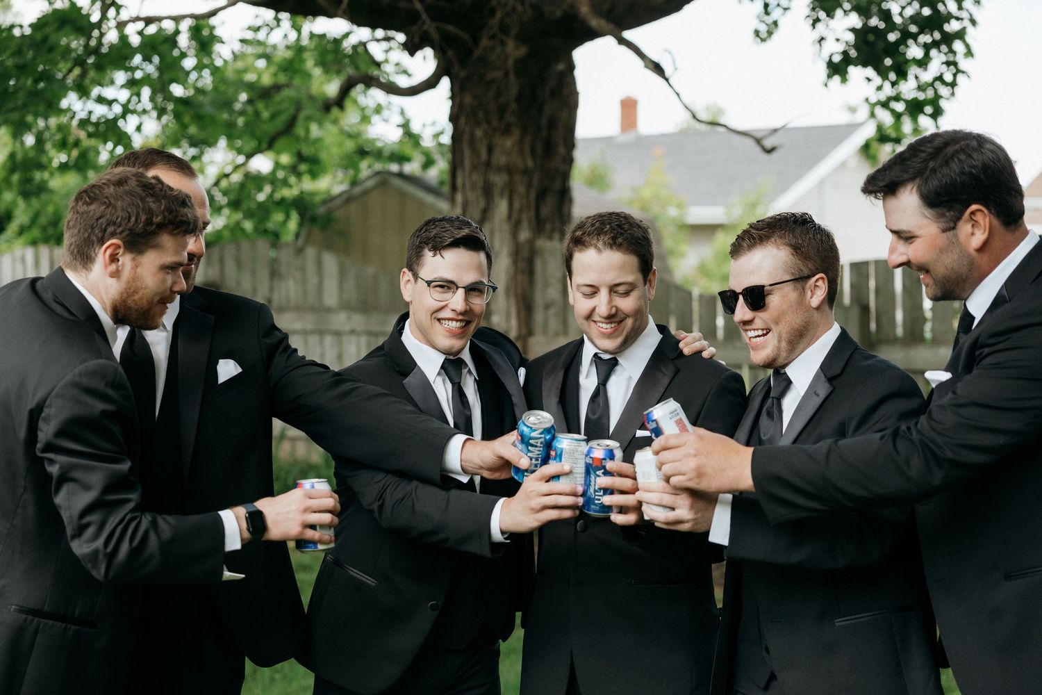 Groomsmen in formal attire share drinks and conversations outdoors.