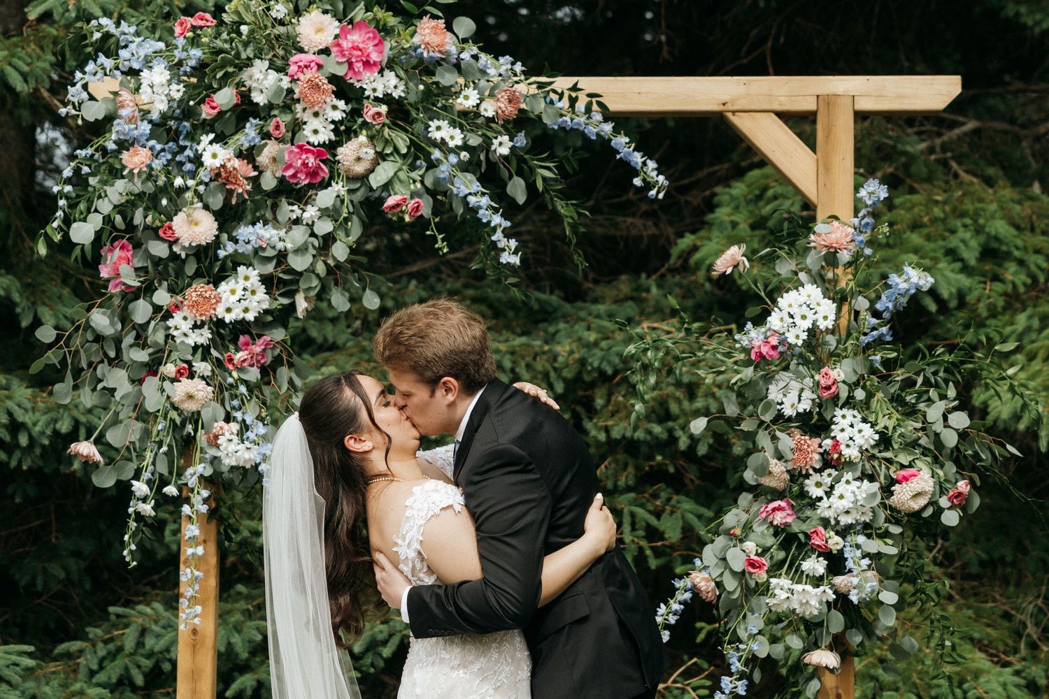 Outdoor wedding ceremony setup with wooden cross arch adorned with pink and white floral arrangements in a garden setting.
