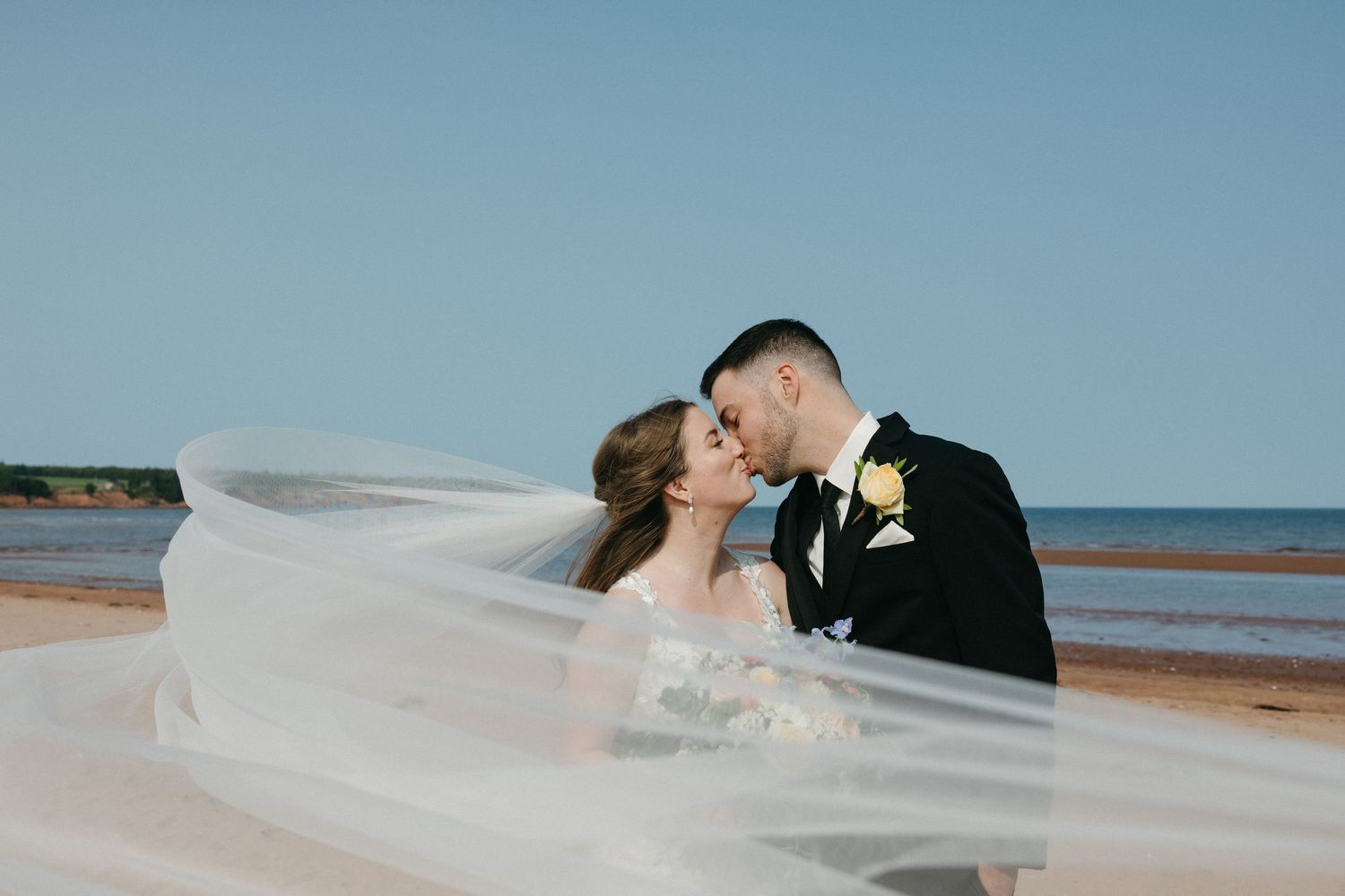 Romantic beach wedding portraits with flowing veil against blue sky.