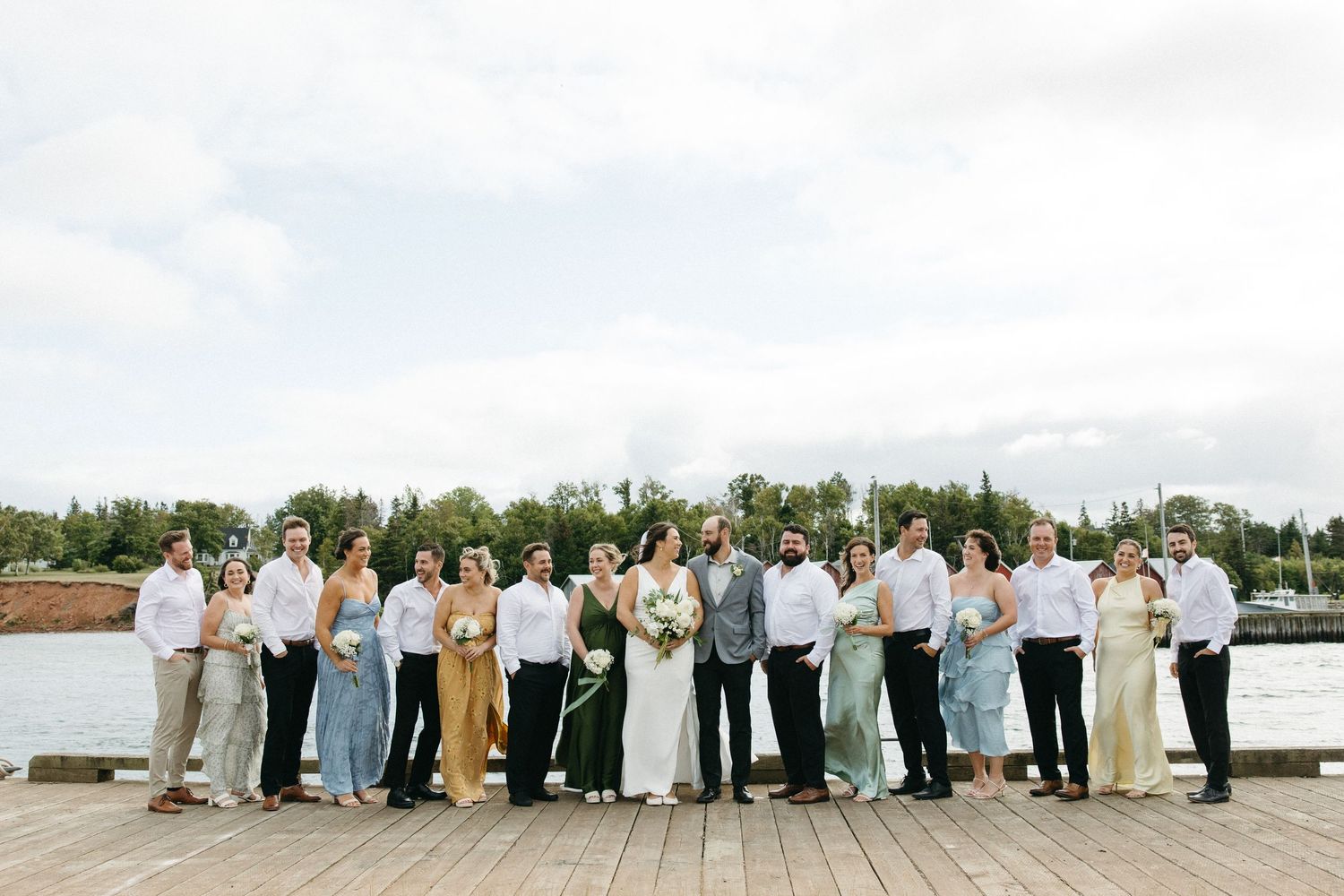 Panoramic shot of wedding guests arranged in line along waterfront with trees in background.