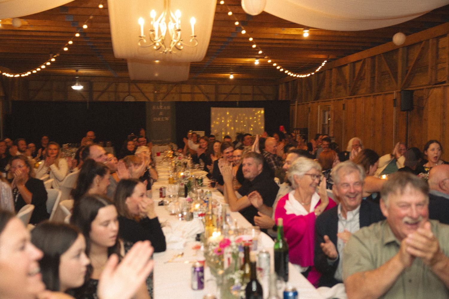 Wedding guests celebrate at reception under chandeliers and string lights in rustic barn setting.