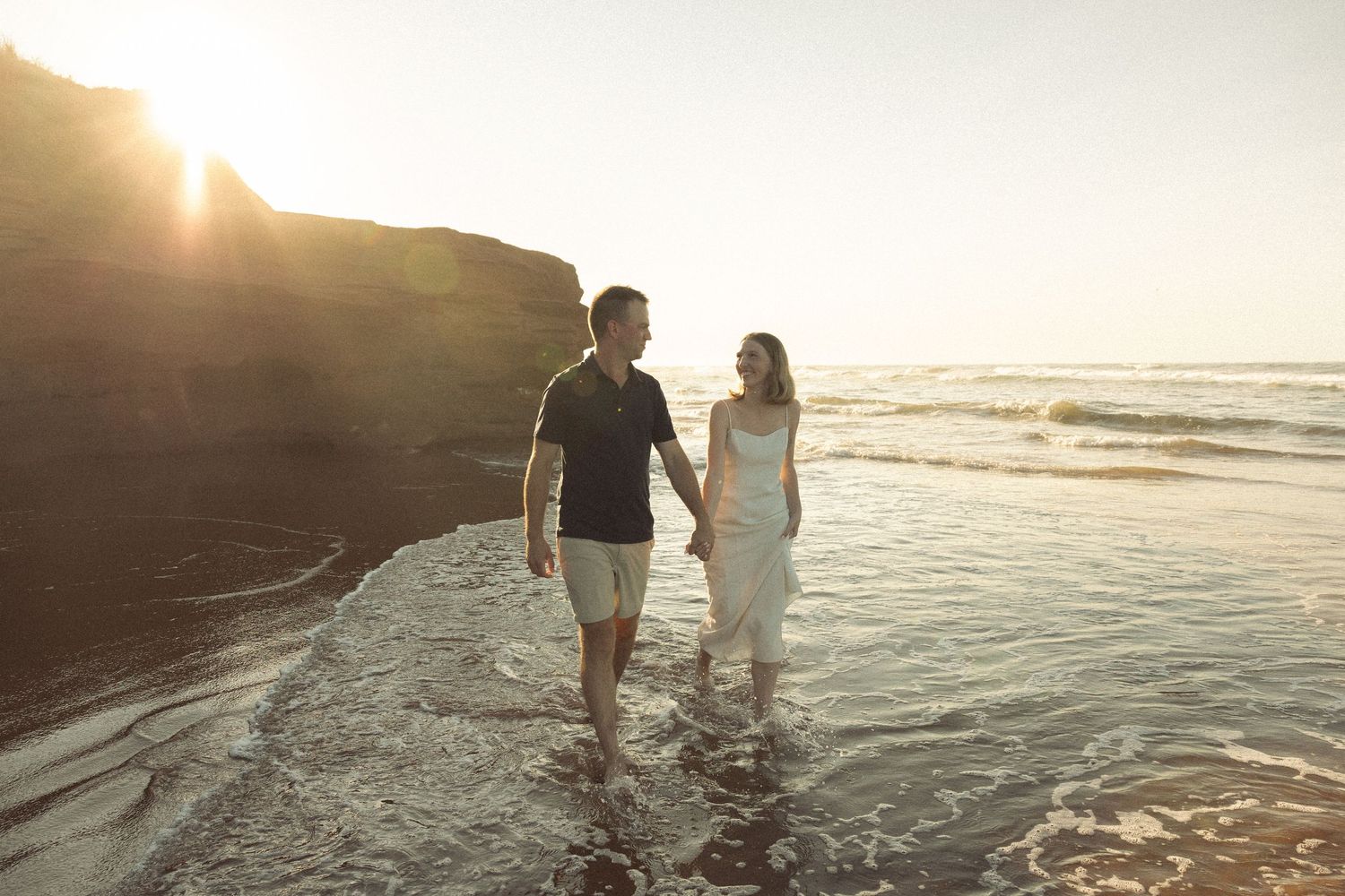 Two people wade through ocean waves at sunset with dramatic coastal cliffs.