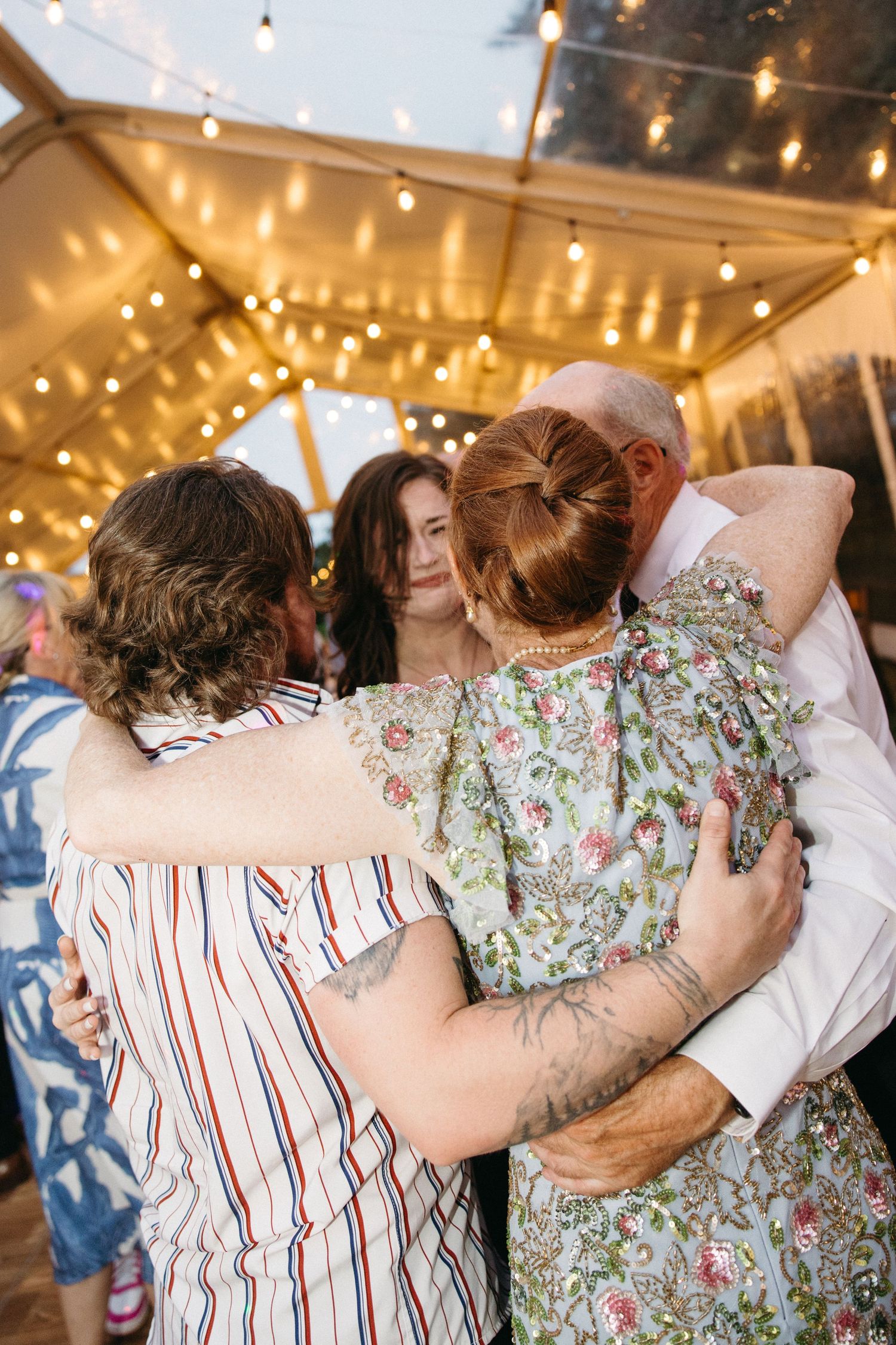 Family members share warm group hug on dance floor under string lights at wedding reception.