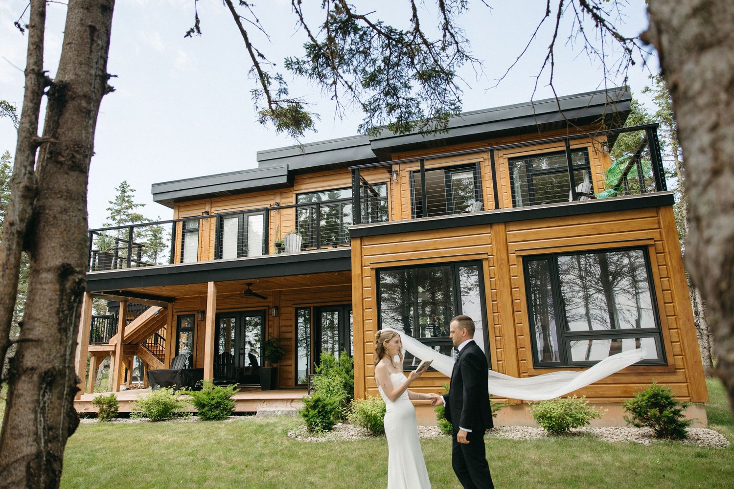 Bride and groom stand on lawn with flowing veil in front of large modern wooden lakeside home.