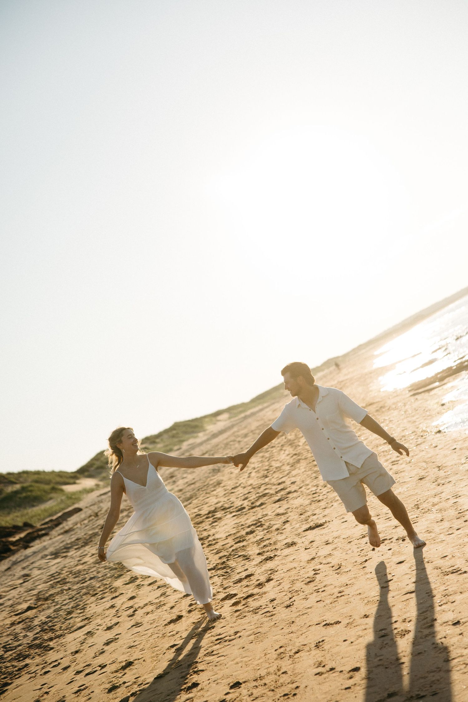 A joyful couple in white run hand-in-hand along a sunny sandy beach, their shadows stretching behind them.