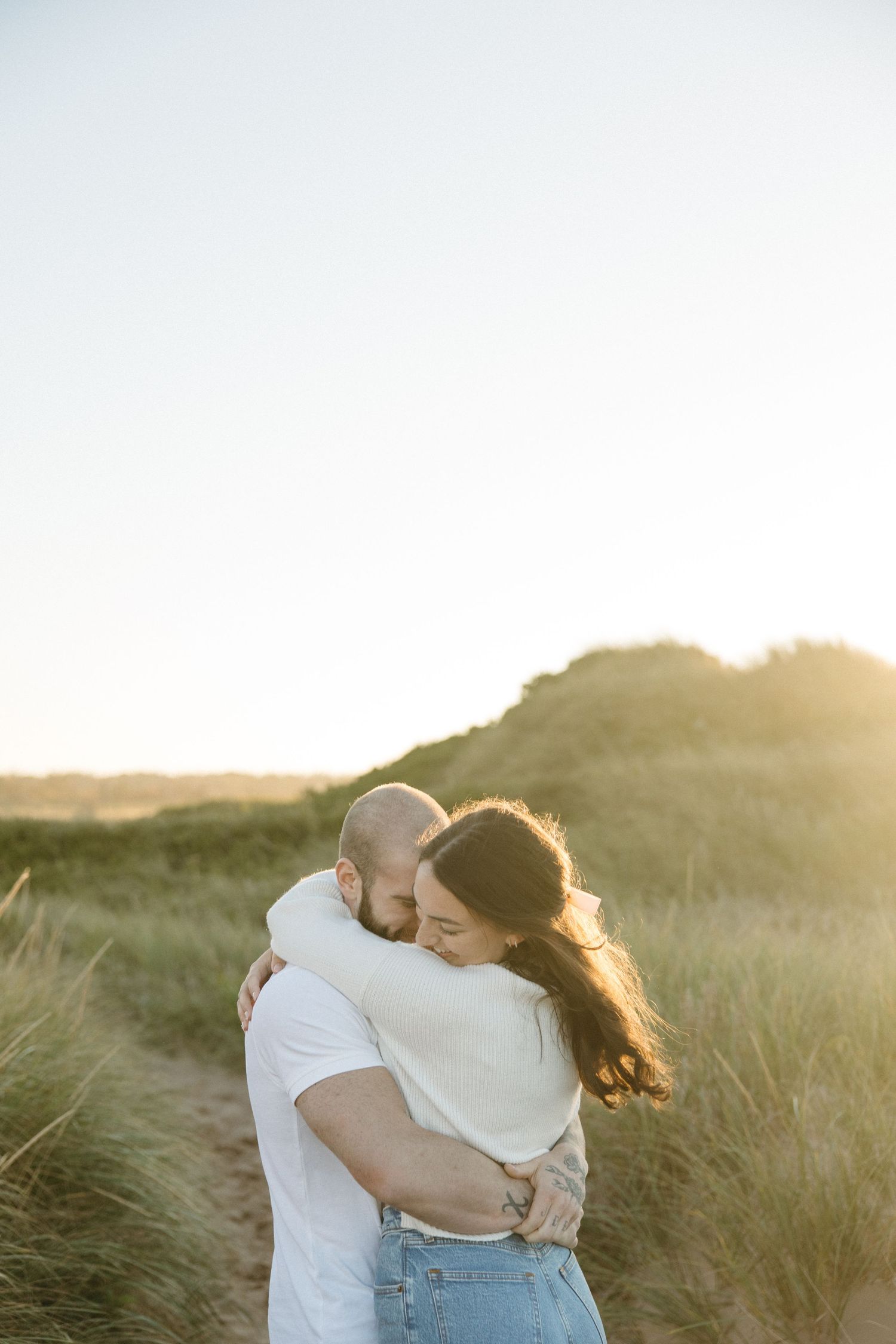 Couple embraces in golden sunlit beach grass during sunset photoshoot.