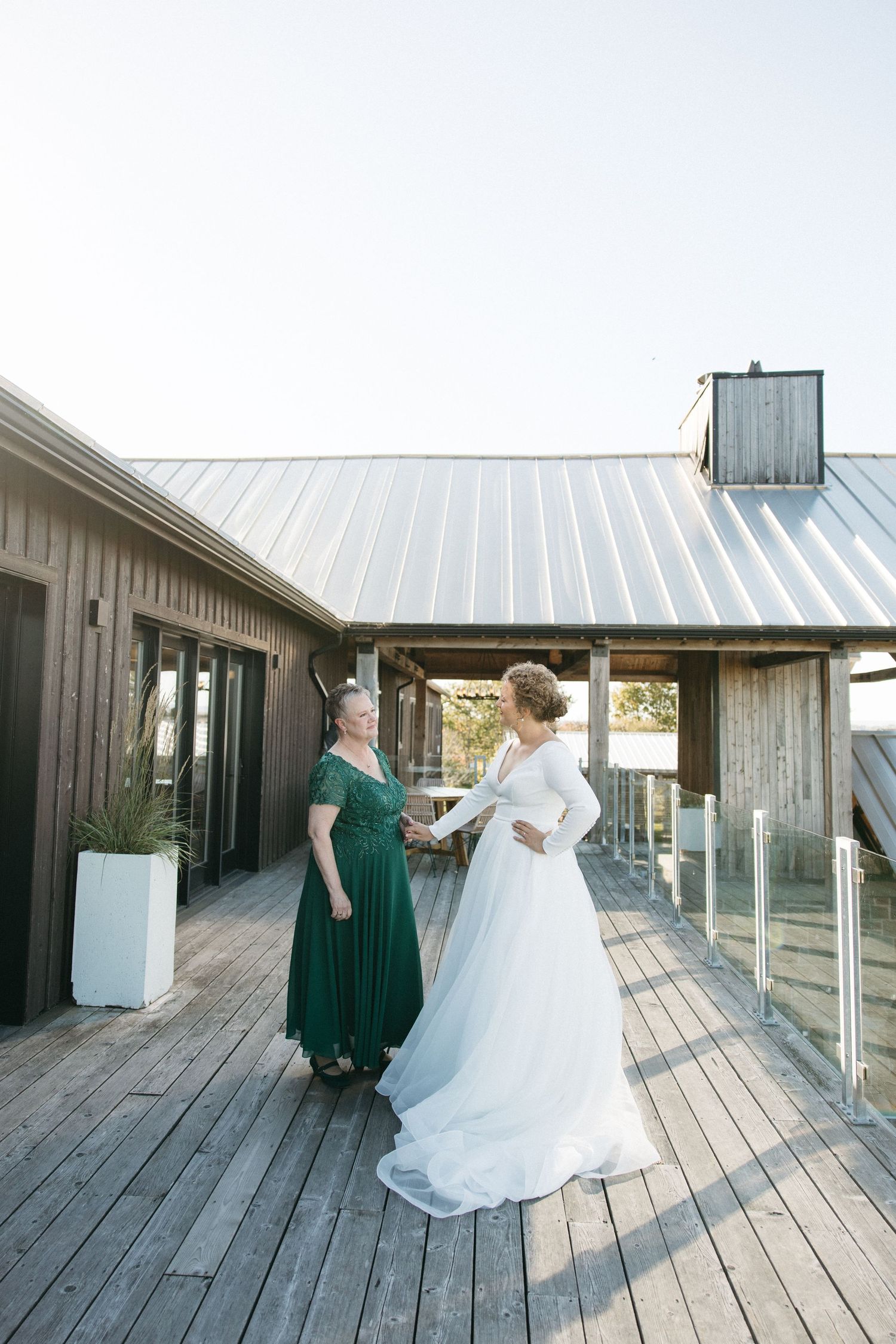 Two people in formal dresses standing on a wooden deck with a modern building exterior.